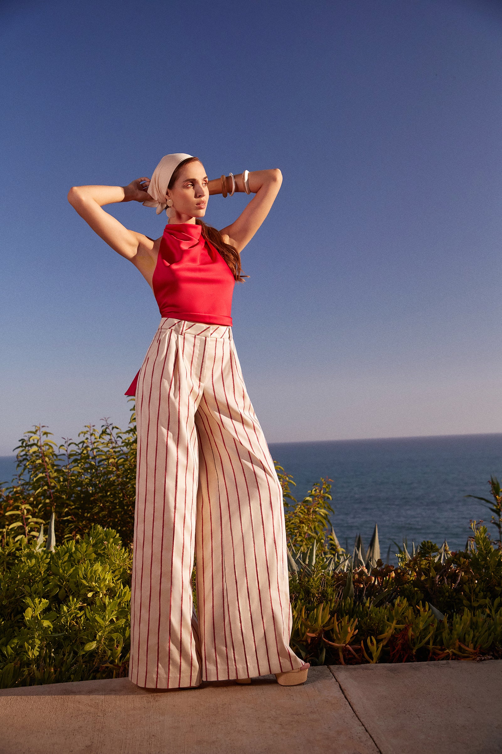 A woman in Solani Wide-Leg Pants - Solaire Stripe stands outdoors by greenery, ocean, and sky, wearing a red sleeveless top and adjusting a white headscarf.