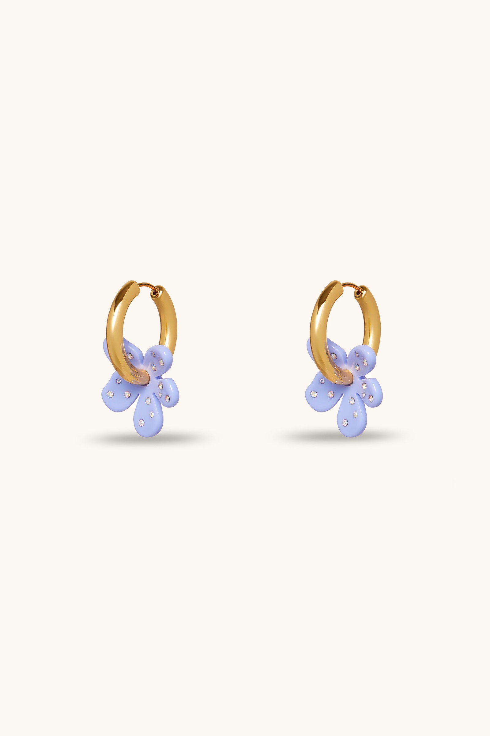 The Lele Sadoughi x Trina Turk Petite Paradis Huggie earrings in periwinkle feature gold hoops with dangling translucent flower charms accented by white dots, shown against a simple light background.