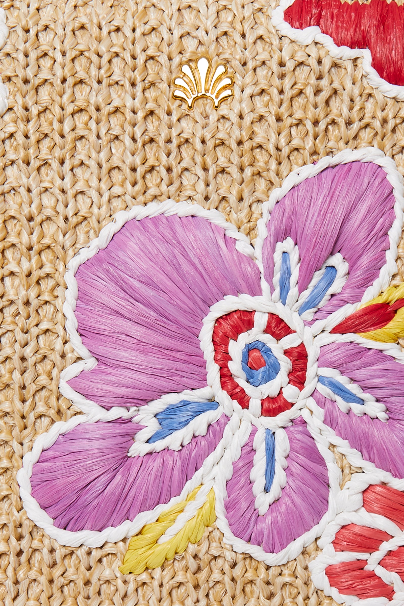 Close-up of the Lele Sadoughi x Trina Turk Maya Floral Mini Tote, featuring a woven beige straw surface with an embroidered purple flower, red and white accents, and a small gold shell emblem above the flower.