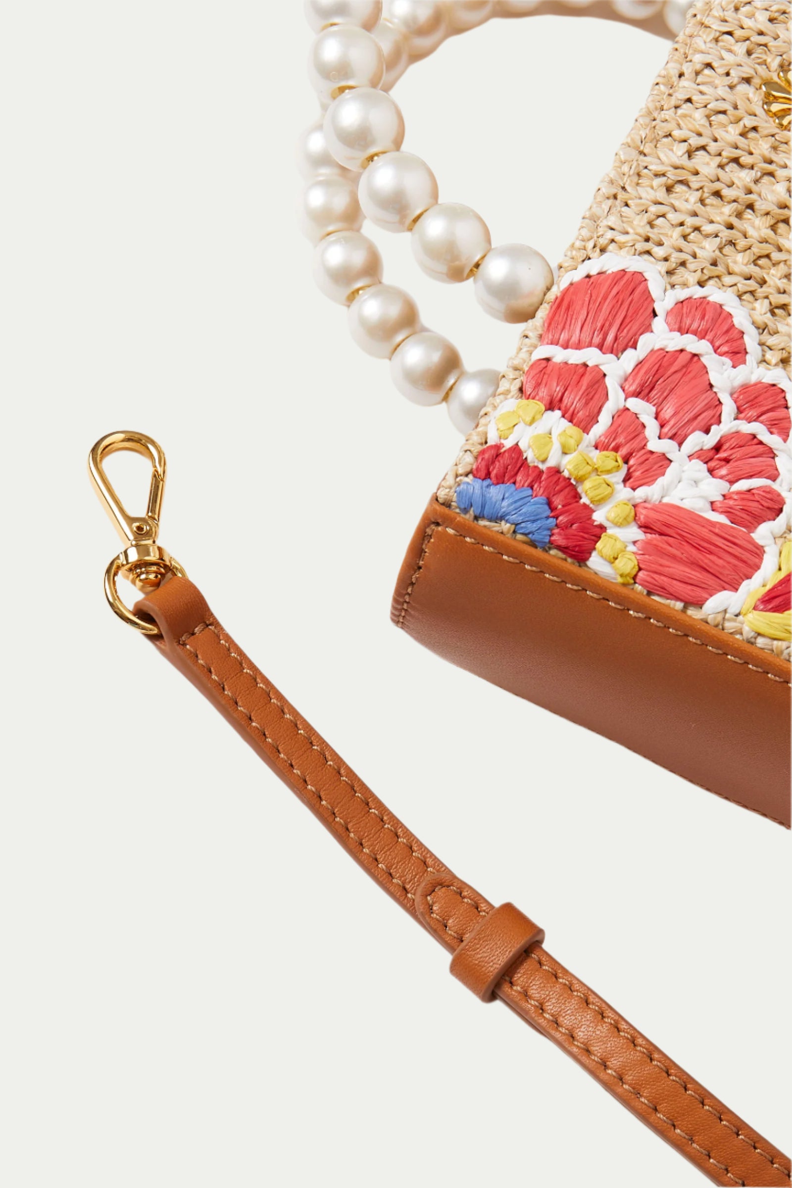 A close-up of the Lele Sadoughi x Trina Turk Maya Floral Mini Tote, featuring brown leather trim, colorful floral embroidery, a detachable leather strap with a gold clasp, and a strand of large white pearls.