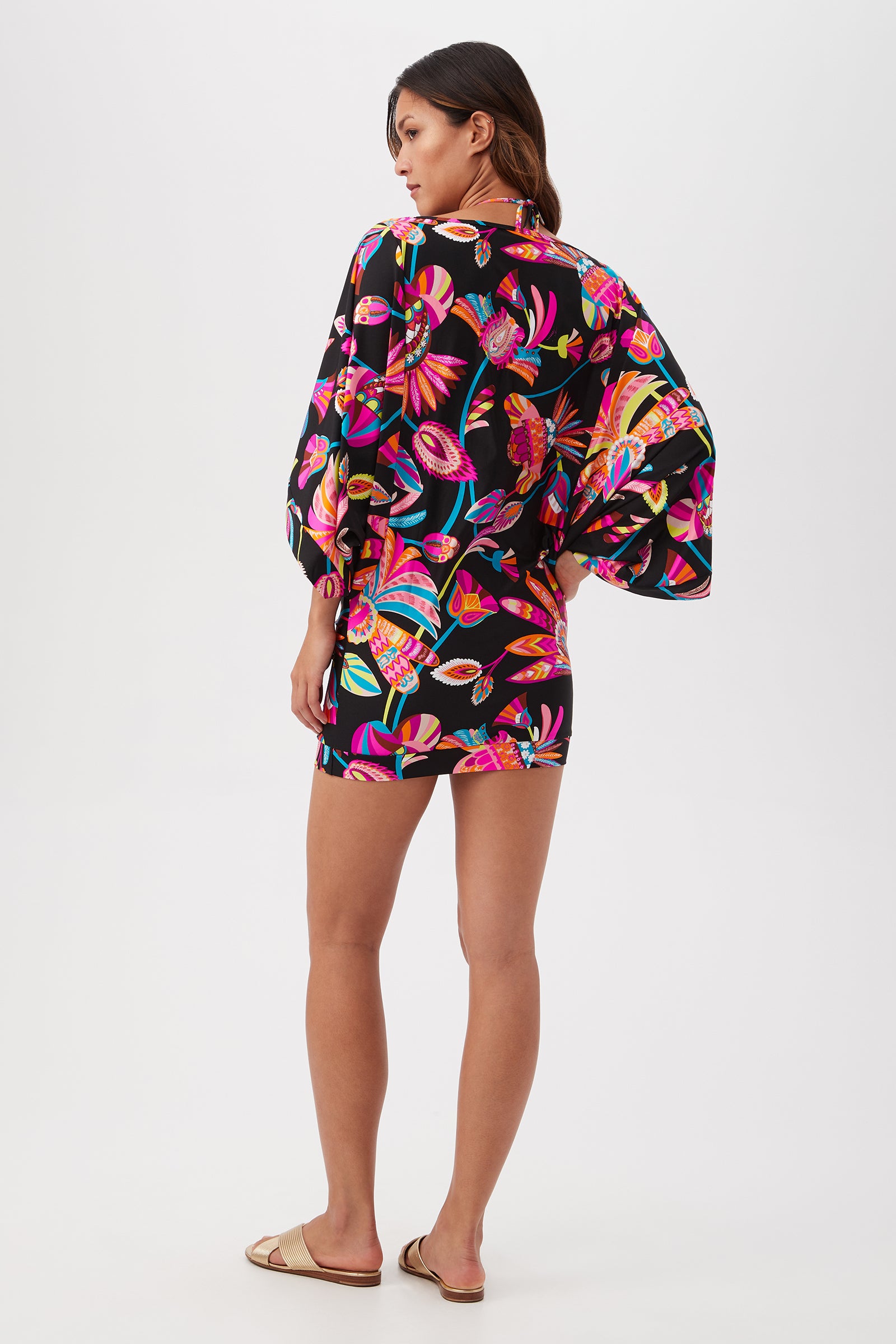 A woman with long hair, in flat sandals, faces away wearing the Luminary Casablanca Tunic Cover-Up—black with vibrant floral and bird prints. Perfect as a swim cover-up for Trina Turk swimwear. Background is plain white.