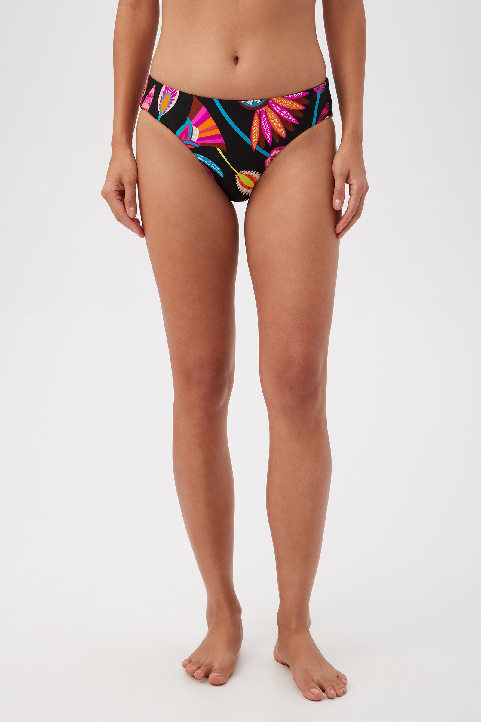 A woman stands barefoot against a white background, wearing the Luminary Reversible Hipster Bikini Bottom in Multi, featuring a bold, colorful floral pattern. Her arms are relaxed at her sides.