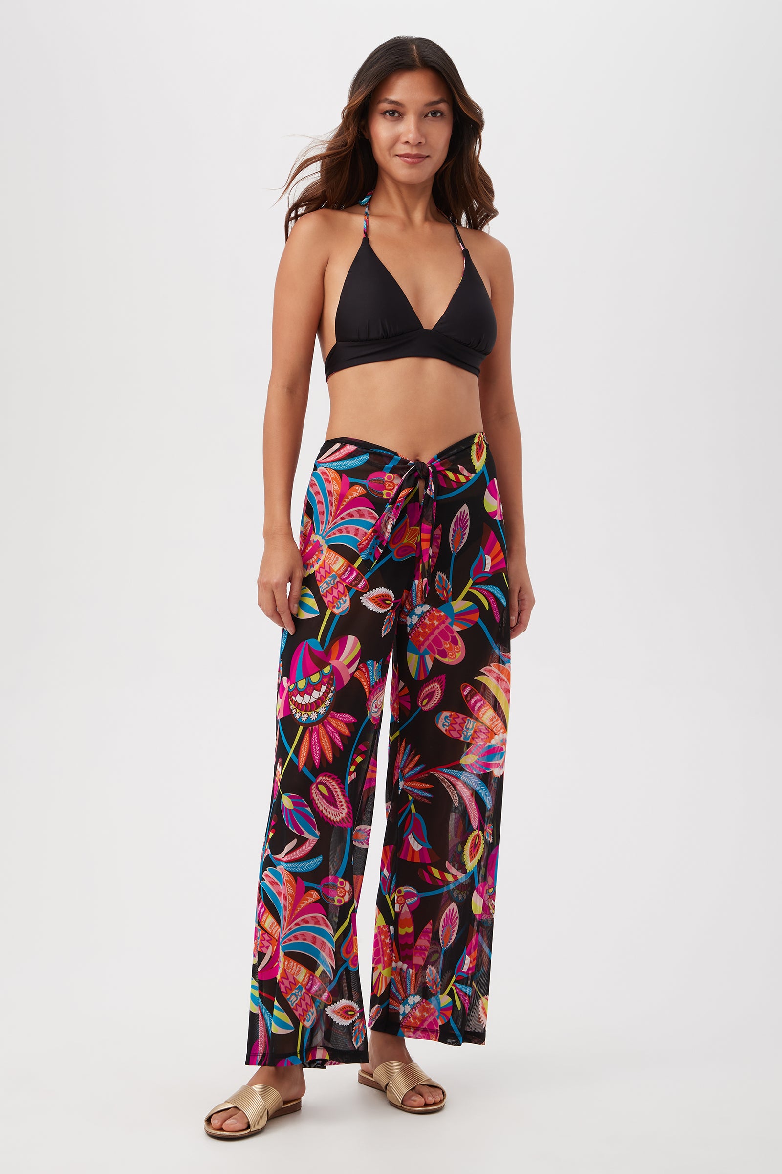 A woman faces forward against a plain white background wearing beige slide sandals, a black bikini top, and the Luminary Mesh Beach Pant Cover-Up featuring a bold tropical pattern.