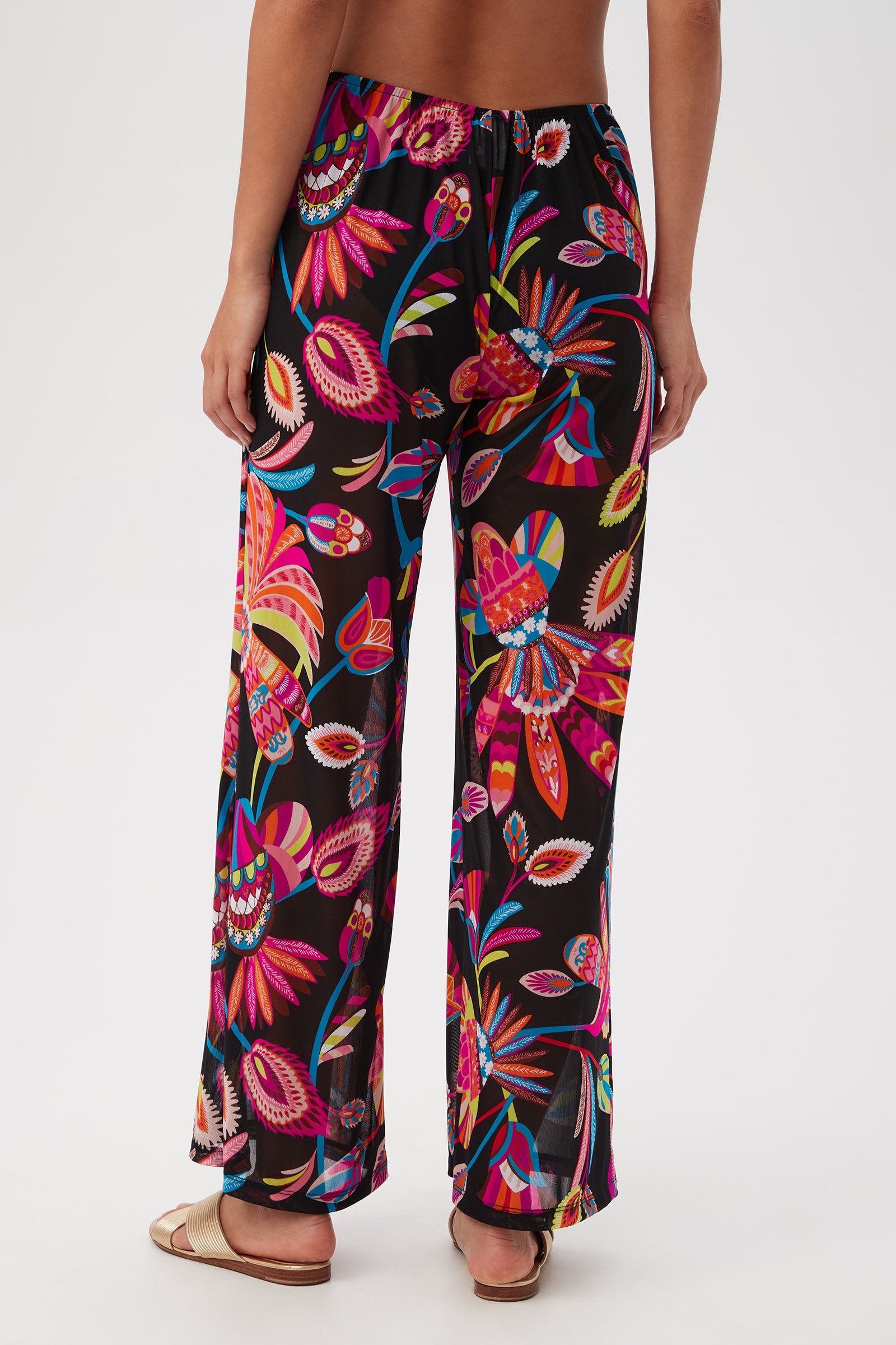 A person stands barefoot on a white background, shown from the back wearing the Luminary Mesh Beach Pant Cover-Up featuring a vibrant tropical pattern with pink, orange, blue, and yellow designs on a black base.