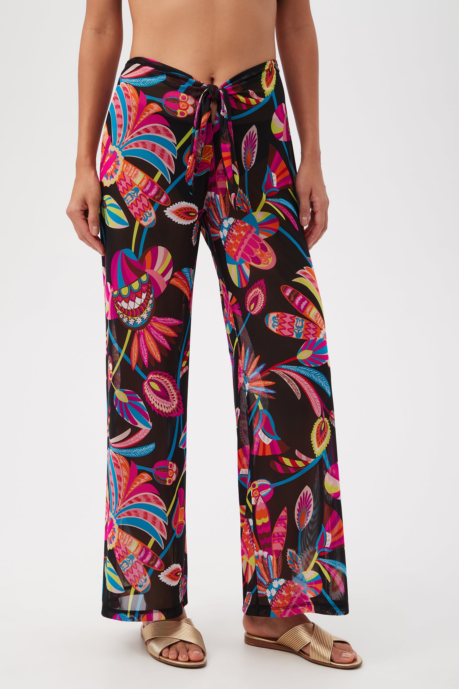A person in Luminary Mesh Beach Pant Cover-Up in Multi, featuring vibrant wide-leg, high-waisted mesh with a tropical pattern, tied at the waist and styled with gold sandals. Upper body is partially out of frame.