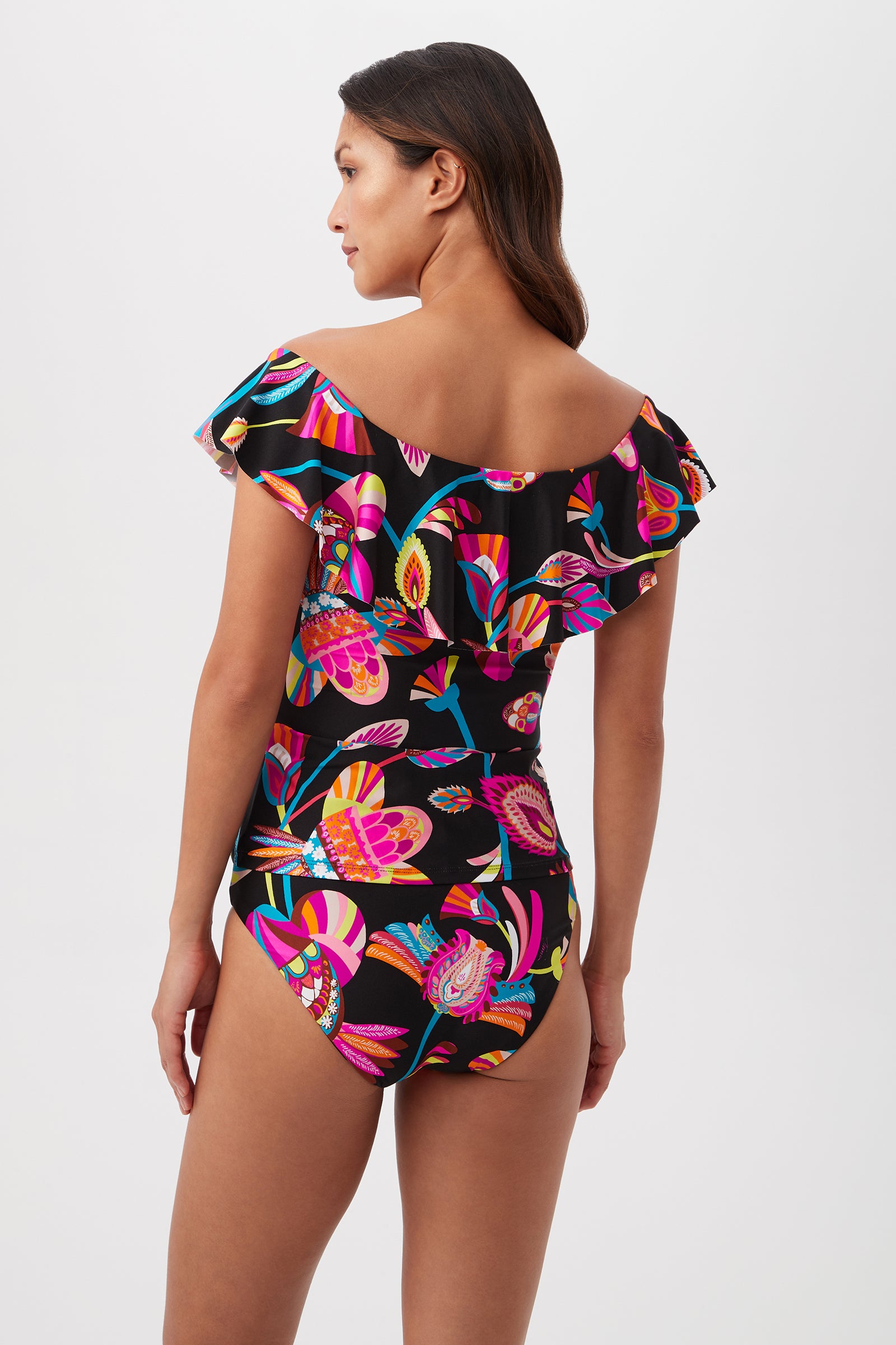 A woman with long brown hair models the Luminary Off-the-Shoulder Ruffle Tankini Swim Top, featuring a vibrant floral and leaf print, shown from the back against a plain white background—an eye-catching swimwear piece.