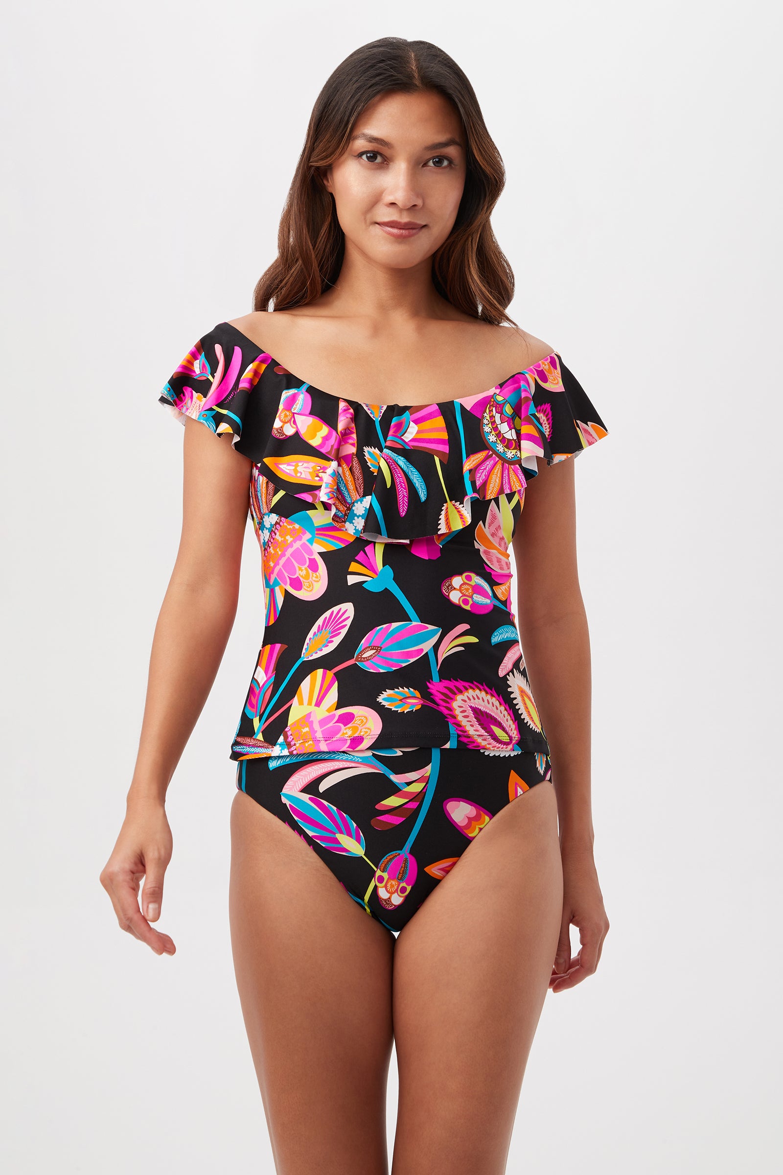 A woman models the Luminary Off-the-Shoulder Ruffle Tankini Swim Top in Multi, featuring vibrant tropical floral patterns and ruffled details, standing against a plain white background.