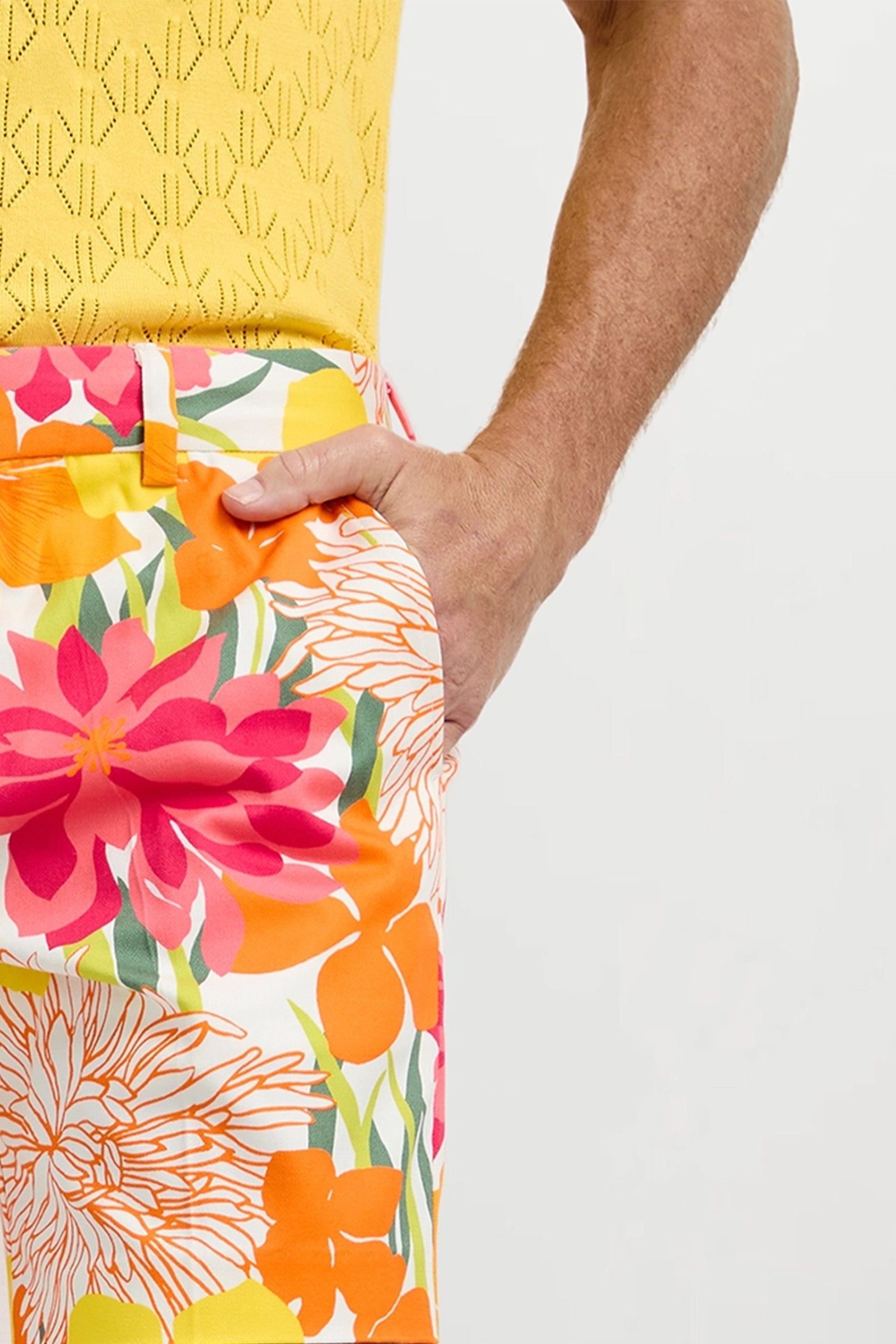 A person models the Lawrence Short - Como Herringbone: yellow textured sleeveless top and vibrant floral pants in orange, pink, yellow, and green, with one hand in a pocket against a plain background.