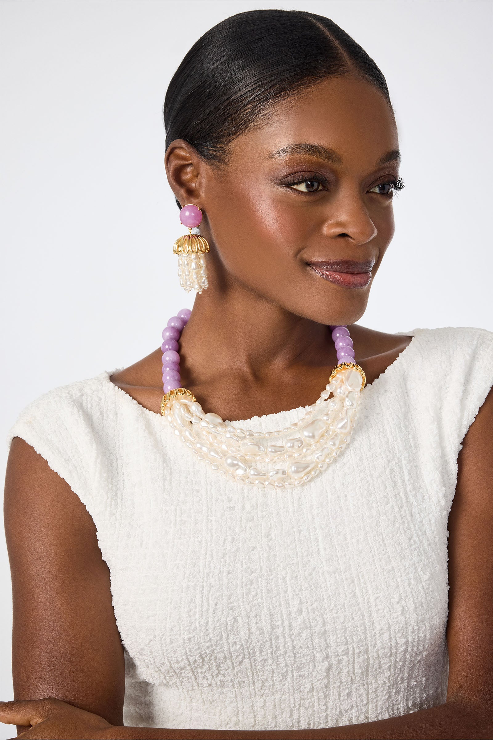 A woman in a textured white sleeveless top wears the Lele Sadoughi x Trina Turk Parapluie Strand Necklace, featuring white and purple beads, along with large gold earrings with purple accents. Her hair is pulled back as she smiles slightly, looking to the side.