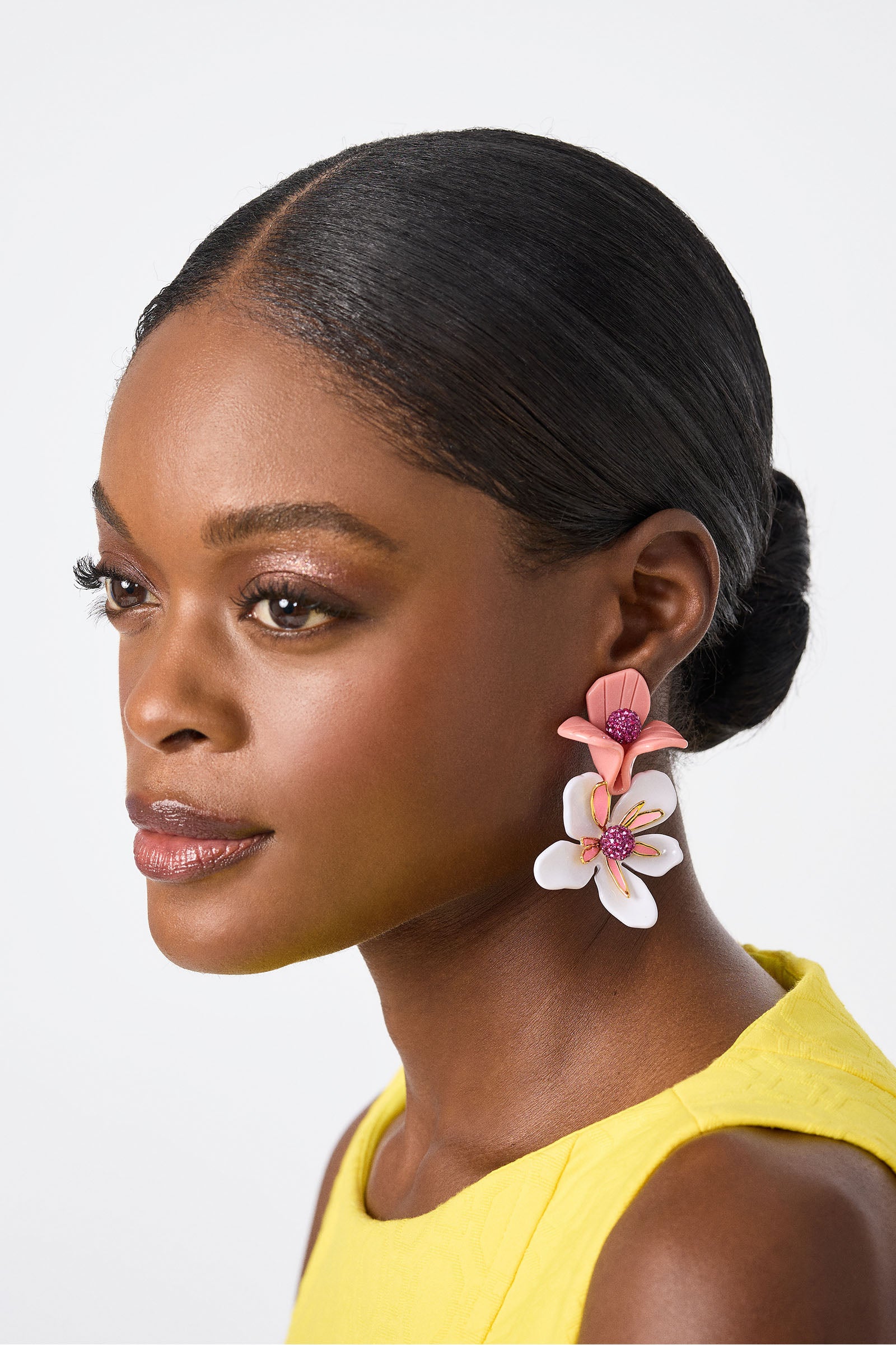 A woman with smooth, dark hair in a low bun wears a sleeveless yellow top and the Lele Sadoughi x Trina Turk Bouquet Paradis Earrings—large, colorful pink and white floral accessories—against a light background, gazing ahead serenely.