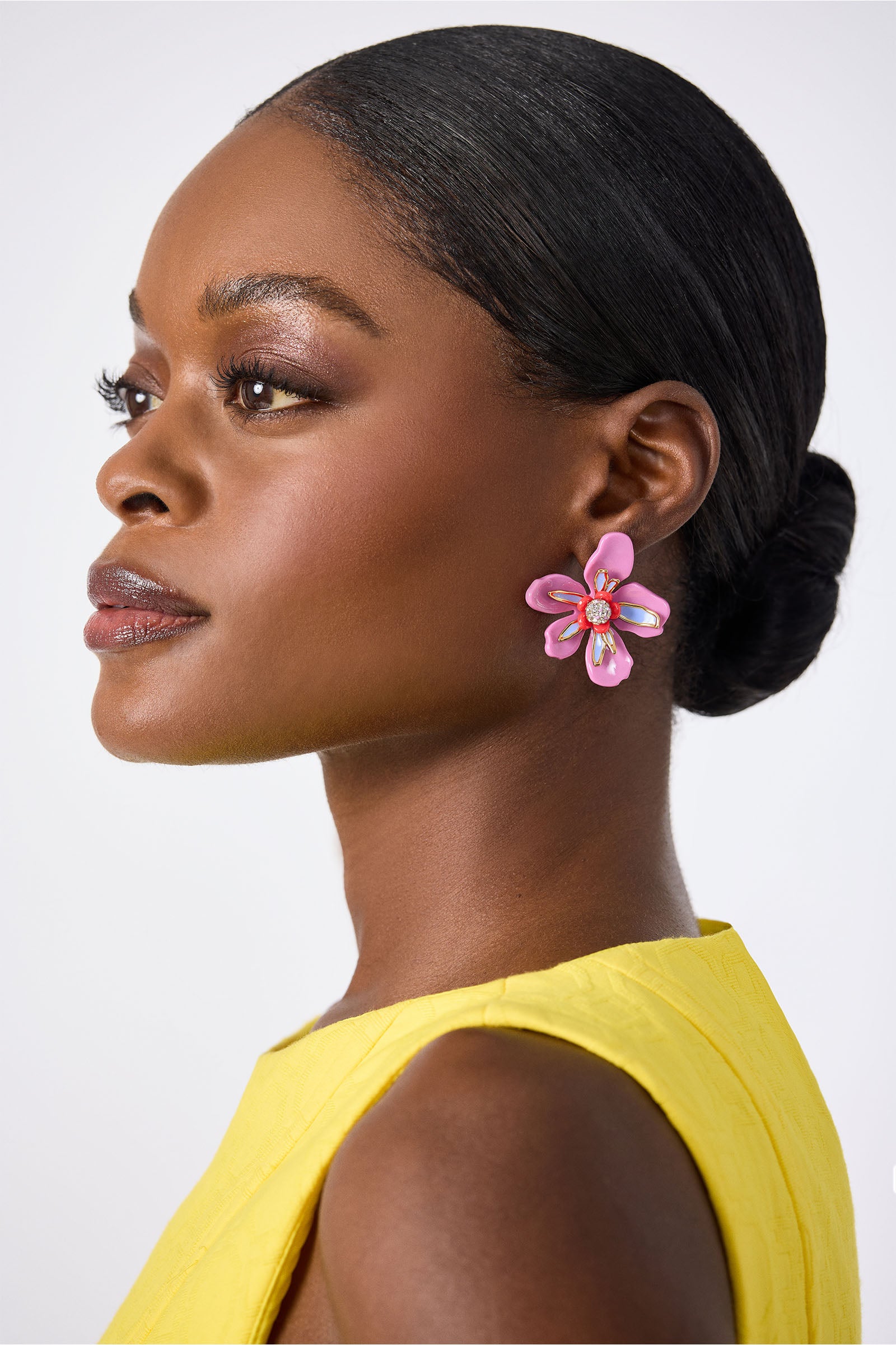 A woman with smooth, dark skin and a low bun wears a sleeveless yellow top and the Lele Sadoughi x Trina Turk Soie Paradis Earrings—large, pink flower-shaped accents—shown in profile against a plain light background.