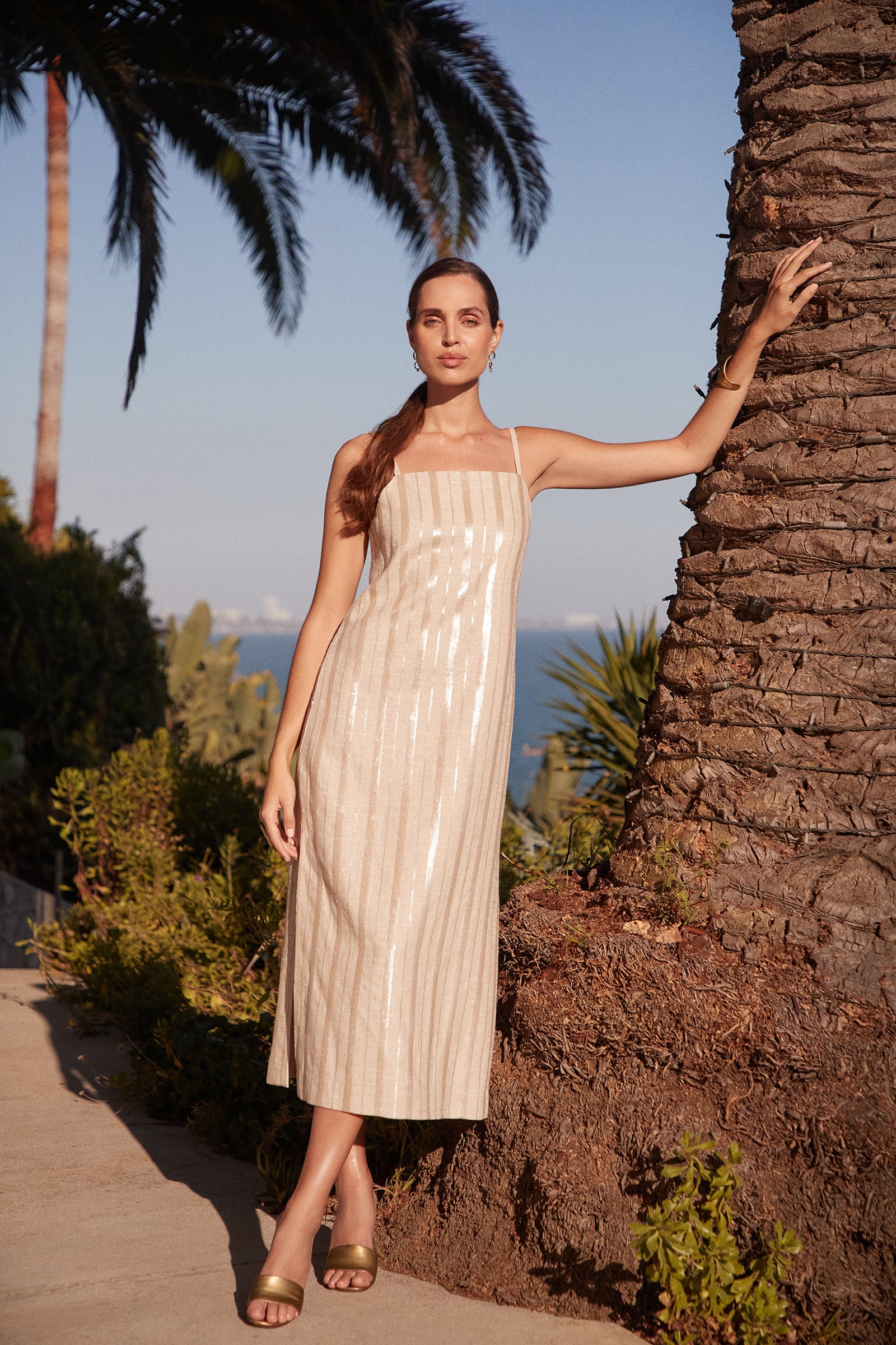 Wearing the Marita 3 Sleeveless Dress in shimmery champagne, a woman stands by a palm tree outdoors, sunlight highlighting her and tropical plants in the background. She rests one hand on the tree and pairs the dress with green sandals.