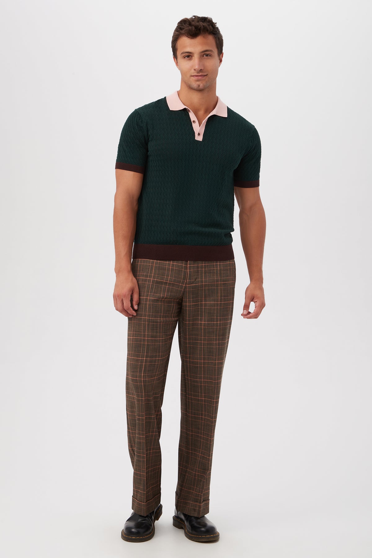 A man stands against a white background, facing forward with a relaxed expression, wearing Genet Wide-Leg Trousers - Chaka Check Plaid, a dark green short-sleeve polo with a pink collar, and black shoes.
