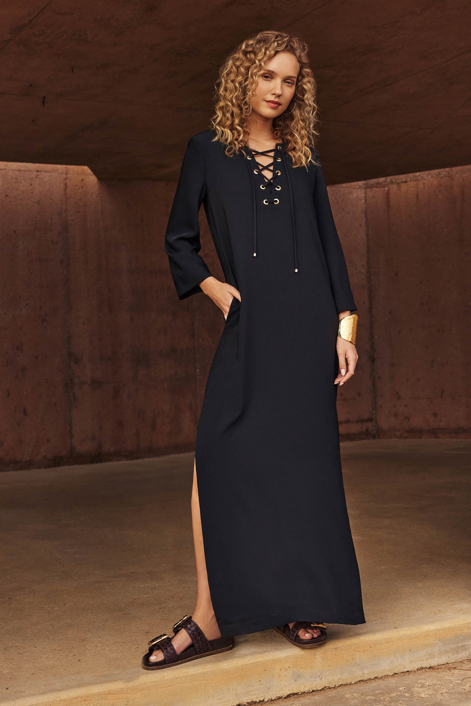 A woman with curly blonde hair wears the Catamaran 3/4-Sleeve Maxi Dress in black, paired with black sandals and a gold cuff bracelet, standing against a brown concrete background.