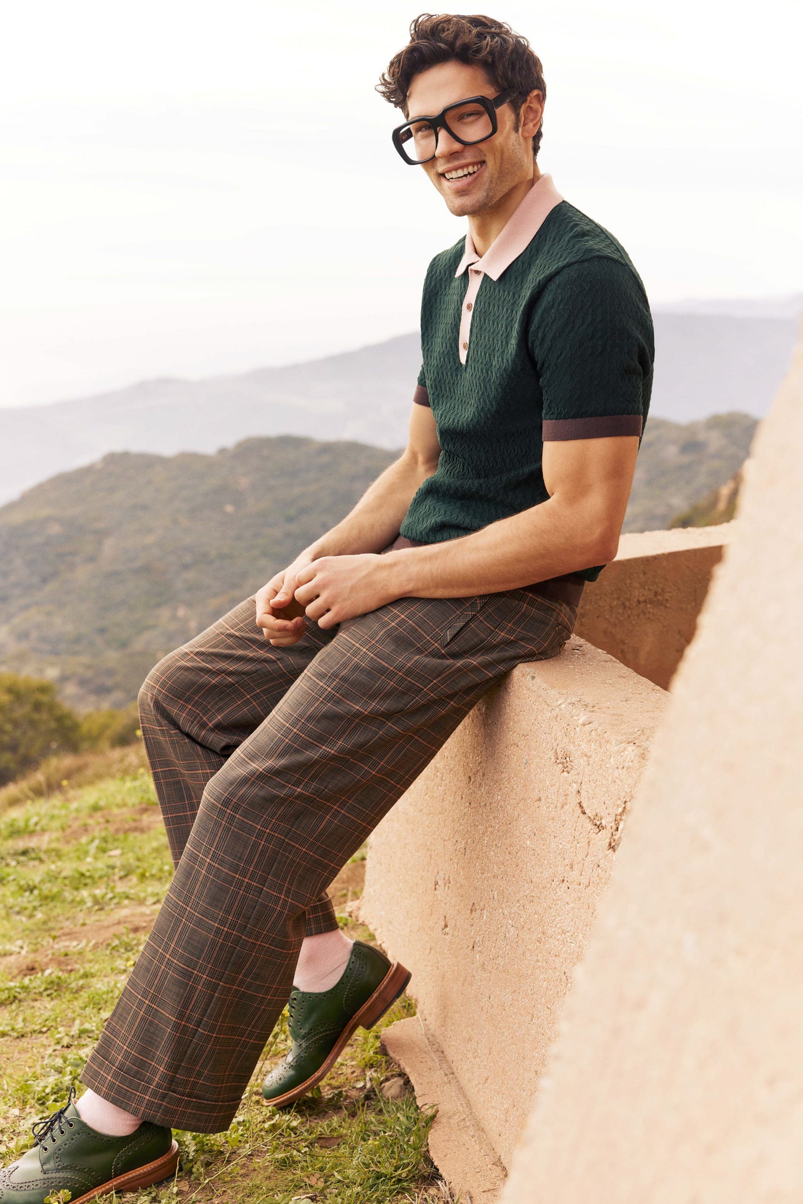 A man with dark curly hair and large glasses sits on a low outdoor wall, smiling. He wears the Vinny Short-Sleeve Polo, plaid brown pants, and green shoes. Mountains and greenery are visible in the background.