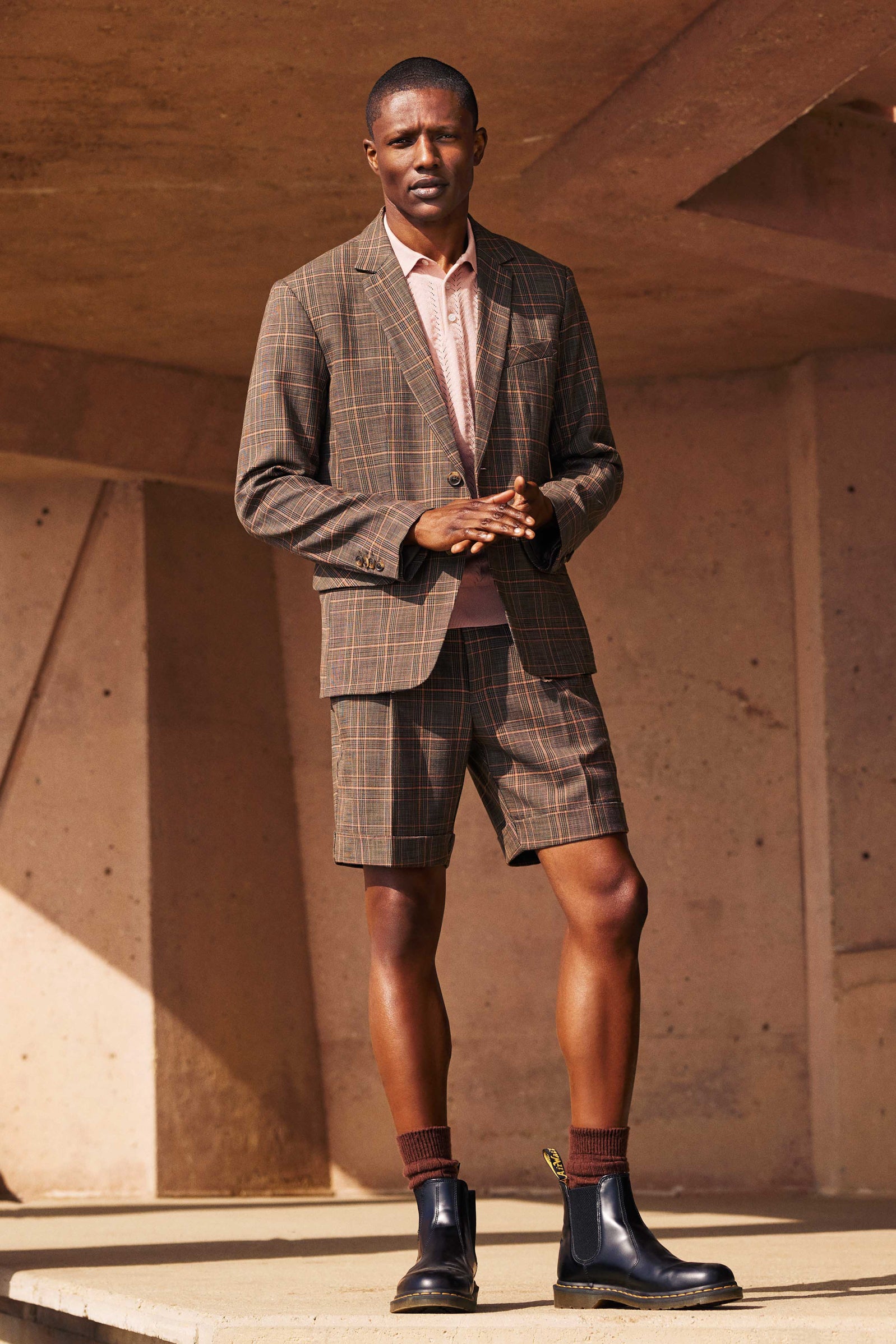 A man stands on concrete wearing the Oliver 2 Short - Chaka Check Plaid, a brown plaid blazer and matching shorts, pink shirt, brown socks, and black ankle boots, set against angular concrete architecture.