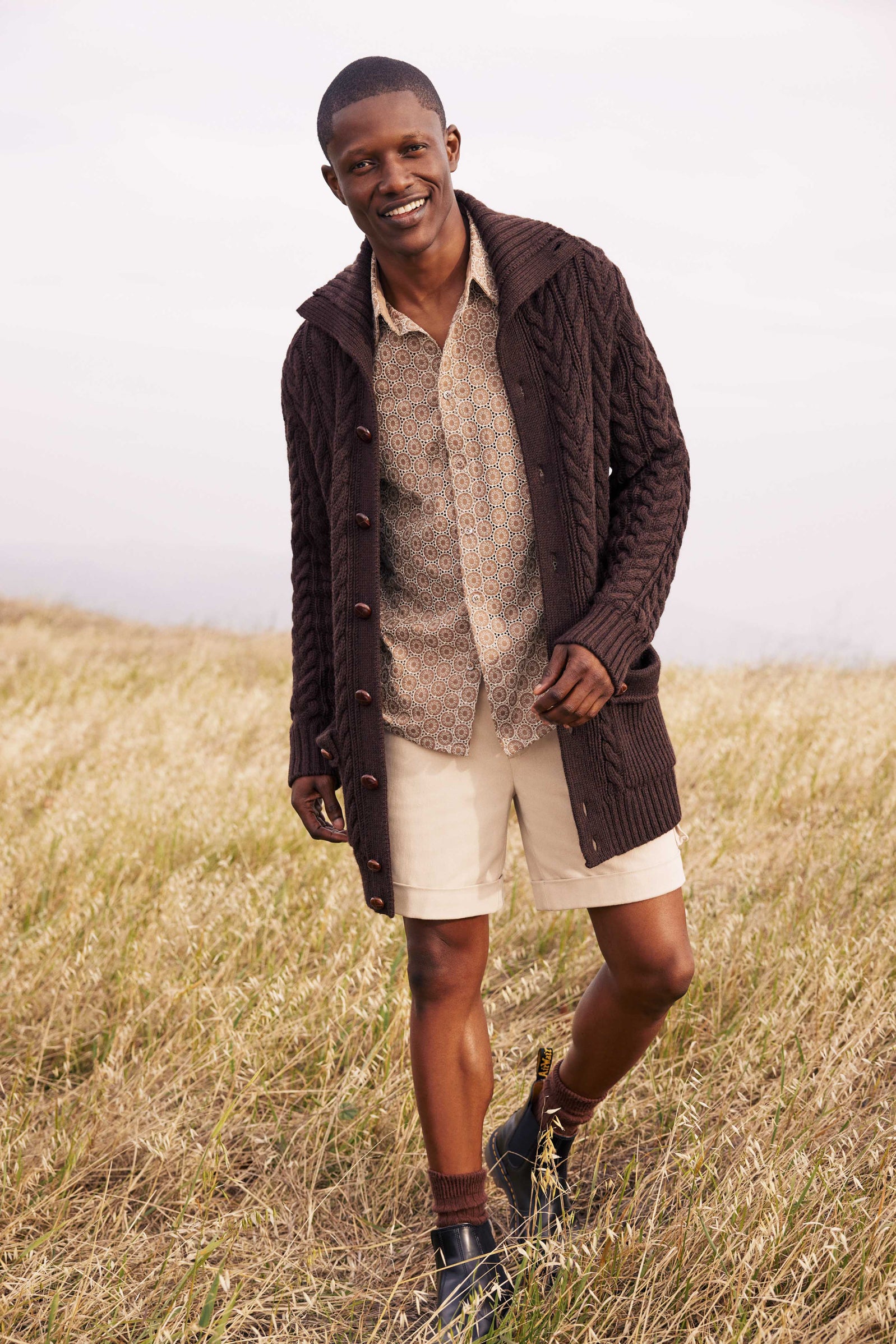 A man stands smiling in a grassy field, wearing the Raleigh Sweater Coat - Merino Wool with a patterned shirt, light shorts, brown socks, and dark shoes.