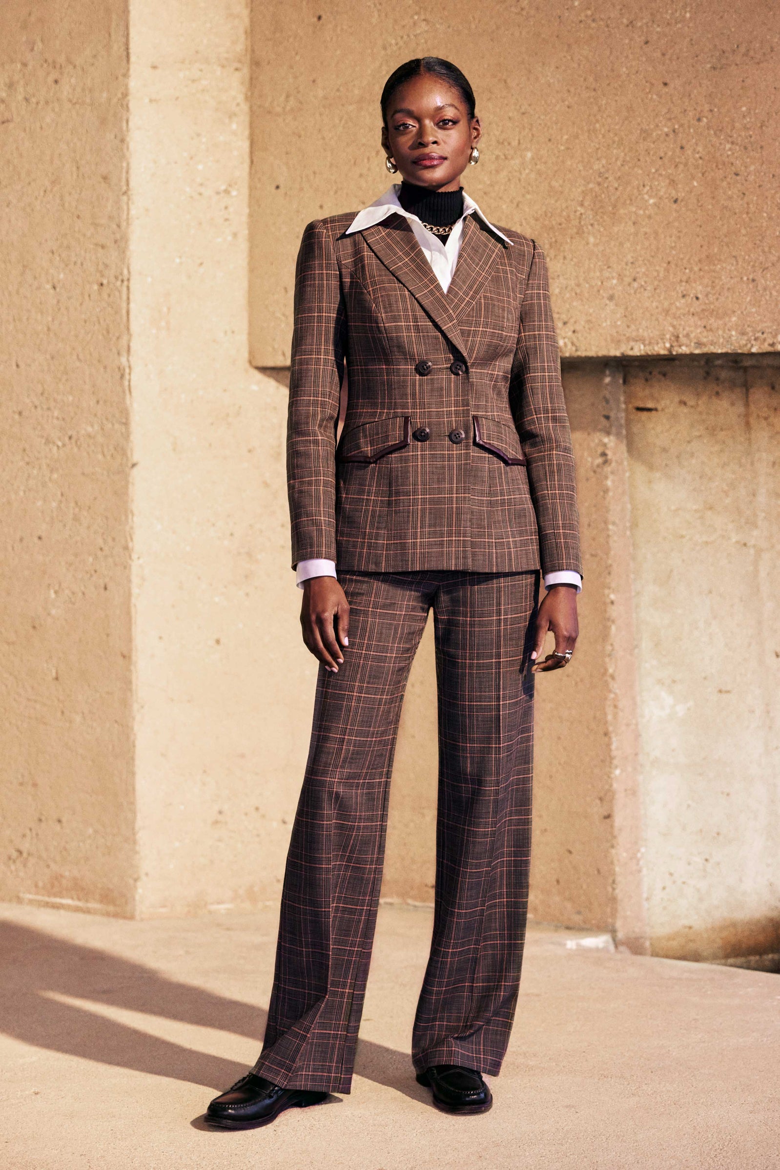 A woman stands confidently against a textured beige wall, wearing the Sadie Pant—a brown plaid double-breasted suit with wide-legged pants—paired with a white collared shirt and black shoes.