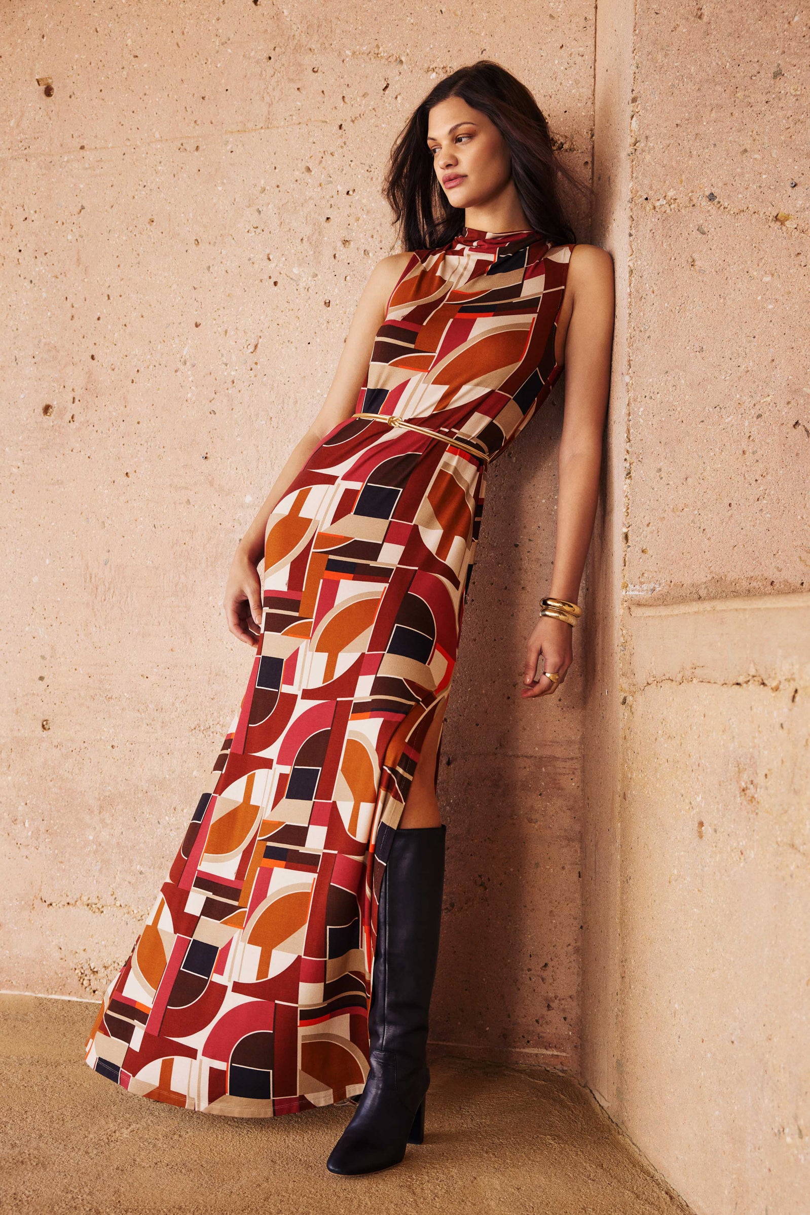 A woman leans against a textured beige wall wearing the Kia Dress, a sleeveless, floor-length piece featuring bold red, orange, black, and cream geometric patterns. She pairs it with knee-high black boots and gold bracelets.