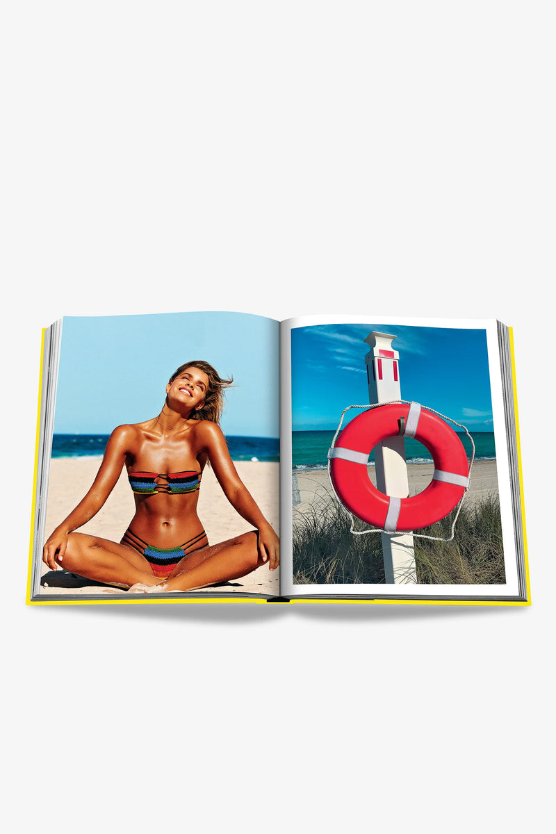 An open Assouline Miami Beach Book displays a woman in a bikini relaxing on the beach on the left page, while the right page features a red lifebuoy mounted on a post by the sea.