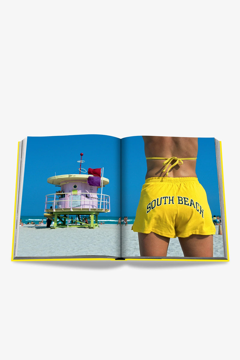 An open Assouline Miami Beach Book displays a beach scene, featuring a colorful lifeguard tower on the left page and a person standing on sand in yellow South Beach shorts on the right page.