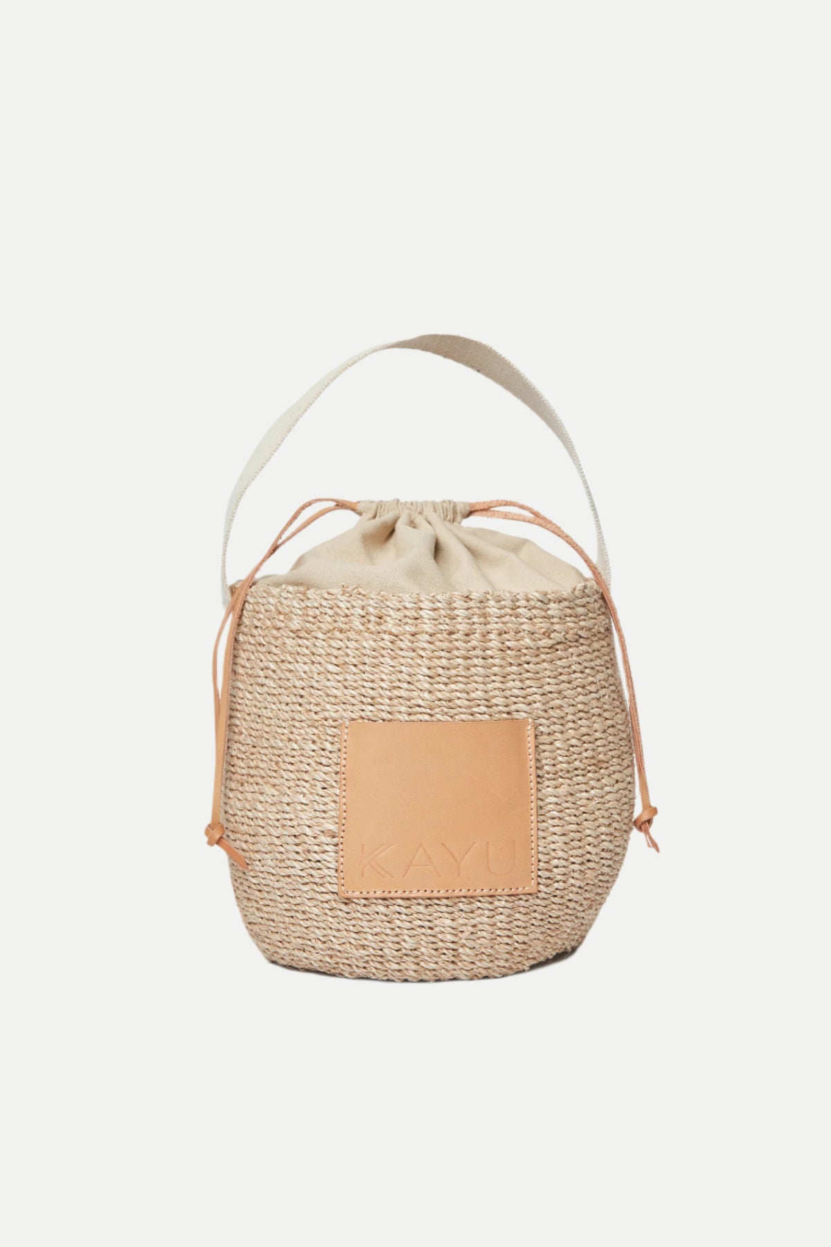 The Mila Straw Bucket Bag features a natural woven straw design, beige fabric drawstring closure, wide handle, and a tan leather KAYU patch on the front.