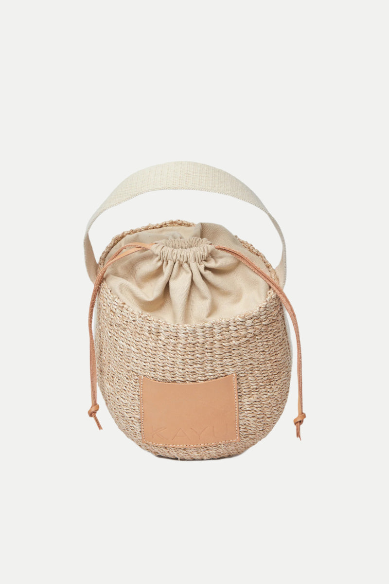 The Mila Straw Bucket Bag features a round woven design with a beige fabric drawstring closure, tan leather accents, and a wide cream shoulder strap, shown against a white background.