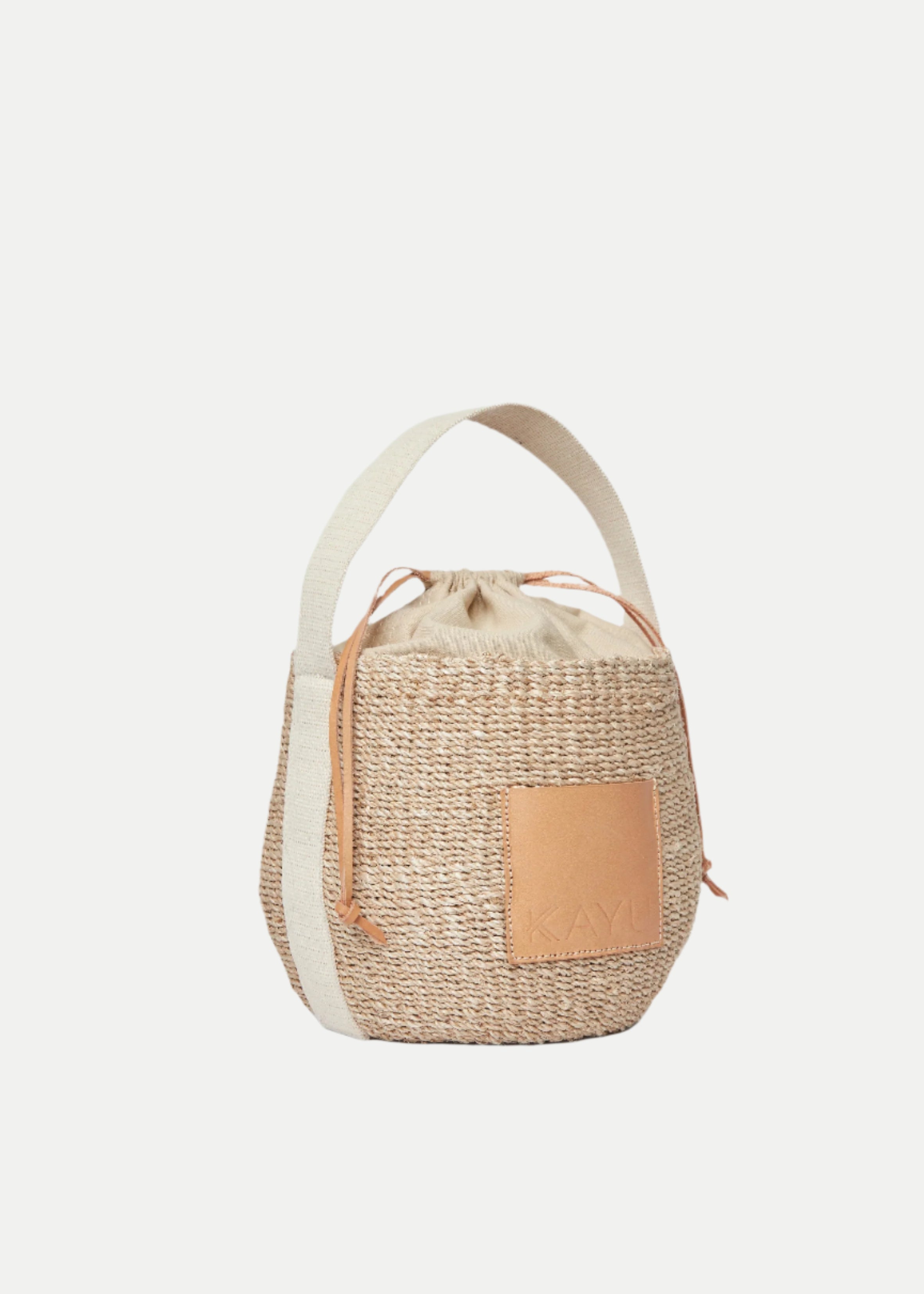 The Mila Straw Bucket Bag features a woven straw design, beige fabric handle, tan leather front patch, and a top drawstring closure.