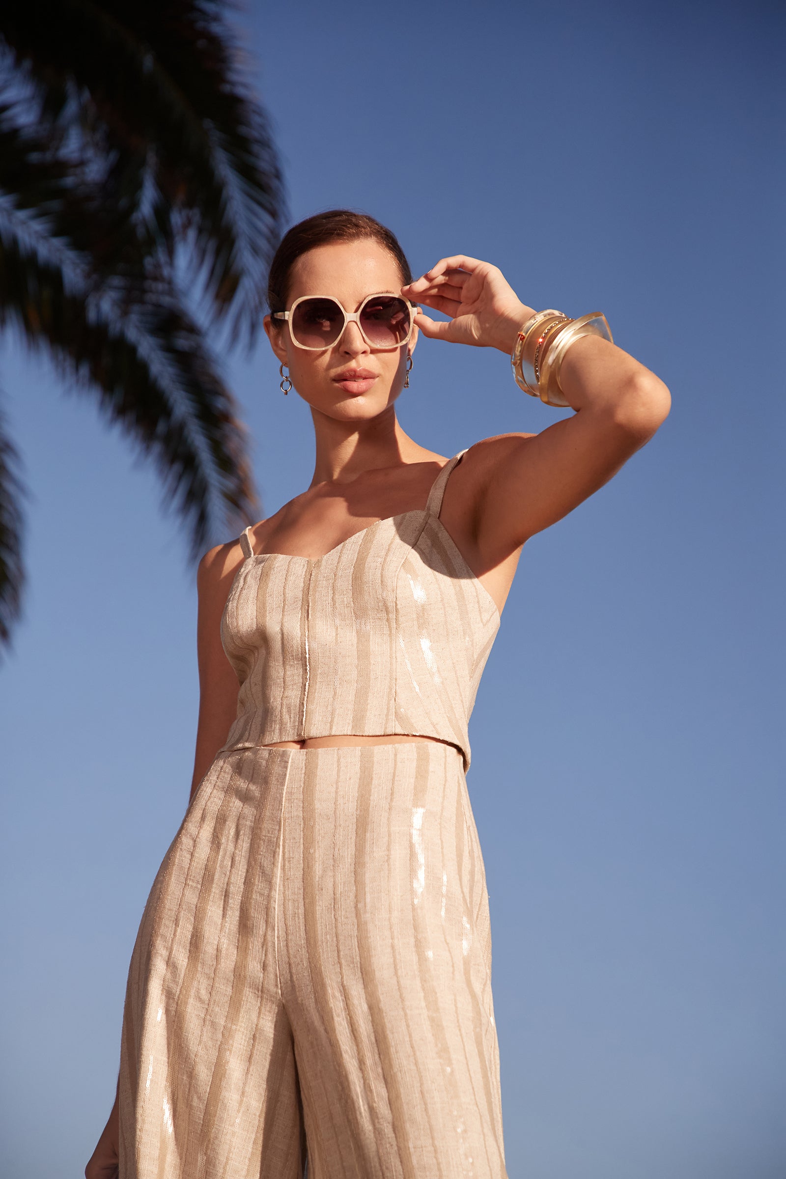 A woman wearing the Millau Sleeveless Top and large sunglasses poses outdoors under a blue sky with a palm tree in the background, accessorized with bangles and holding one hand near her sunglasses.
