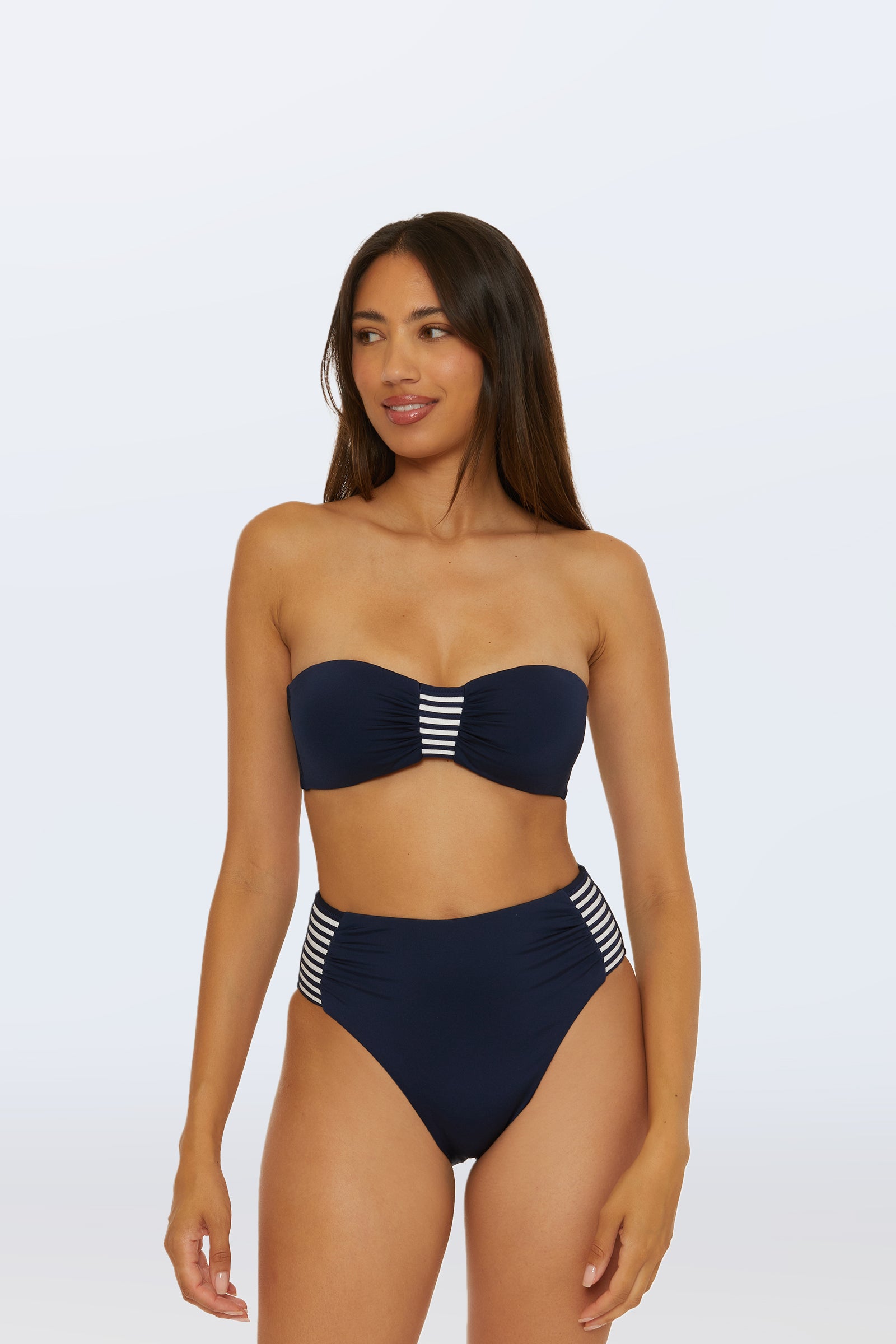 A woman wearing the Monaco Pleated Tab Bandeau Swim Top in Ink, featuring pleated detailing and striped accents, stands against a plain light background.