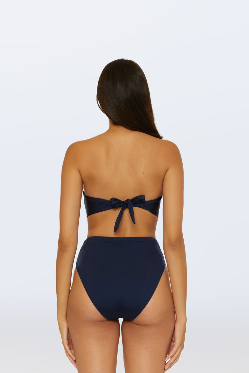A woman with long brown hair is shown from behind, wearing the Monaco Pleated Tab Bandeau Swim Top in navy blue with a high-waisted bottom. The bandeau top features a bow tie at the back against a plain light gray background.