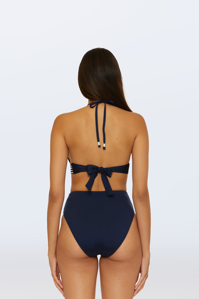 A woman with long brown hair is shown from the back wearing the Monaco Pleated Tab Bandeau Swim Top in navy blue, paired with matching high-waisted bikini bottoms, set against a plain light background.