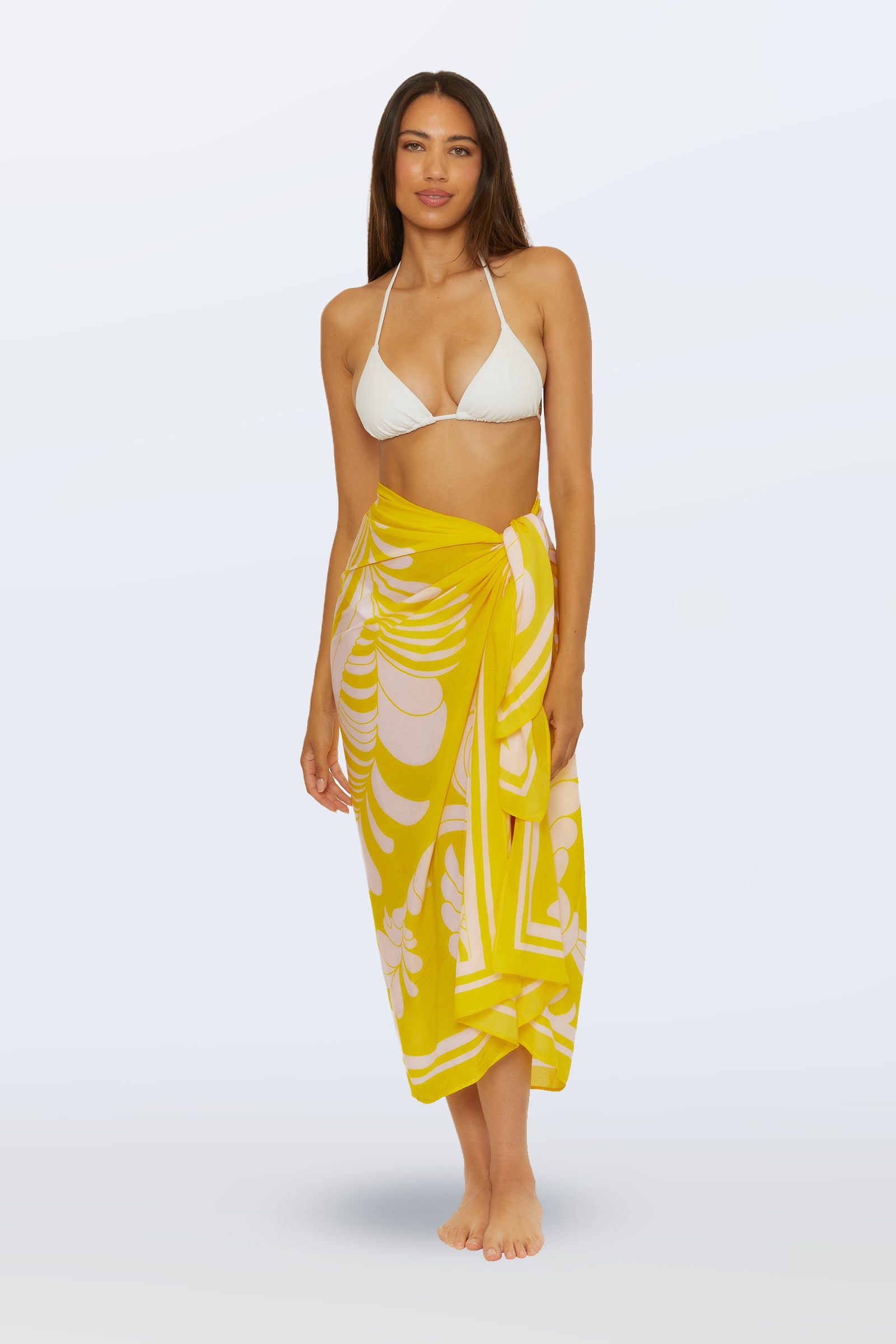 Woman stands barefoot against a plain light background, smiling and wearing a white bikini top paired with the Pareo Swim Cover-Up in Desert Sun (Monaco), featuring white tropical leaf patterns.