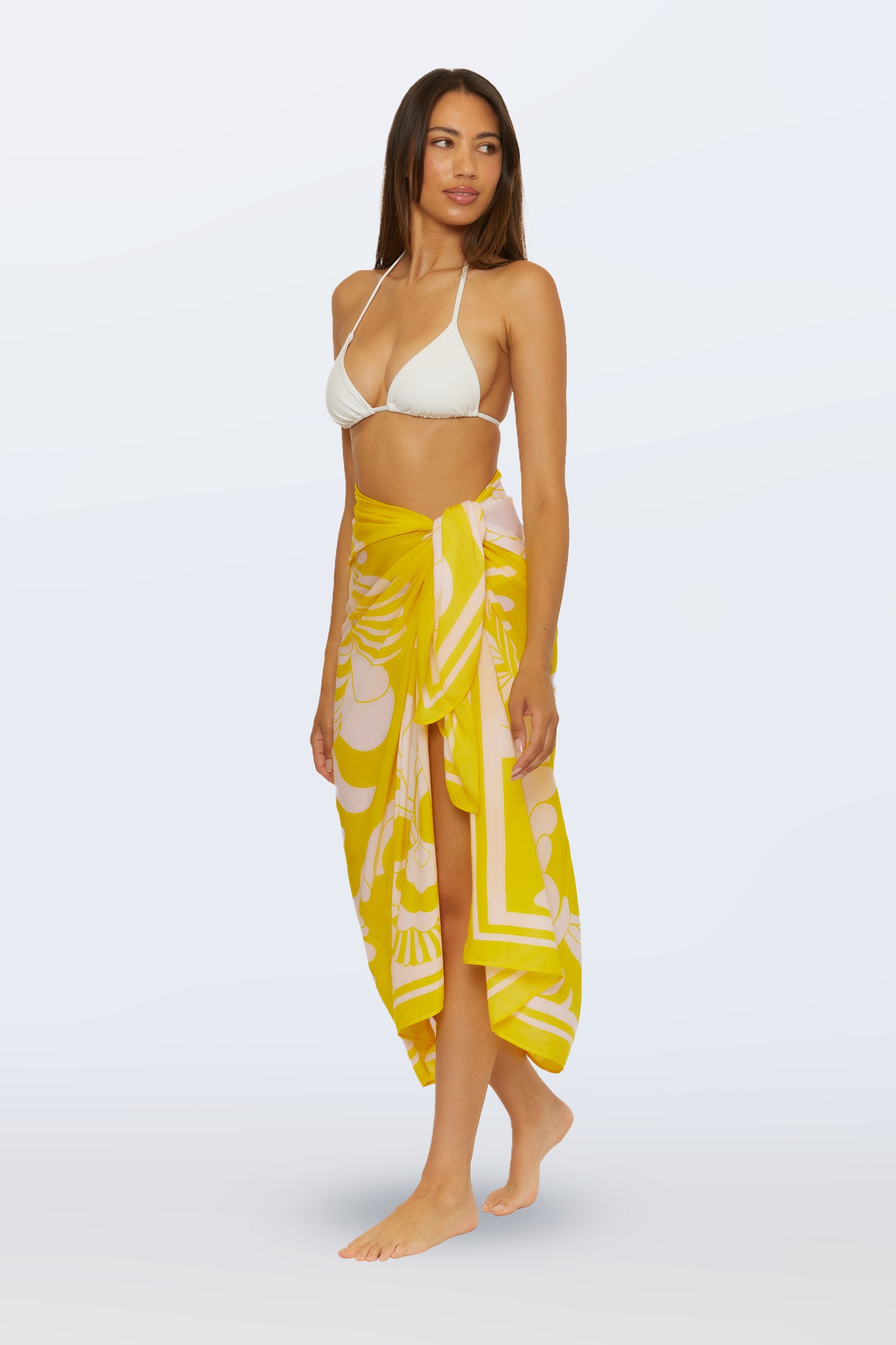 A woman stands barefoot against a light background, wearing a white bikini top and the Pareo Swim Cover-Up – Monaco Desert Sun, a yellow sarong with a white floral pattern tied at her waist.