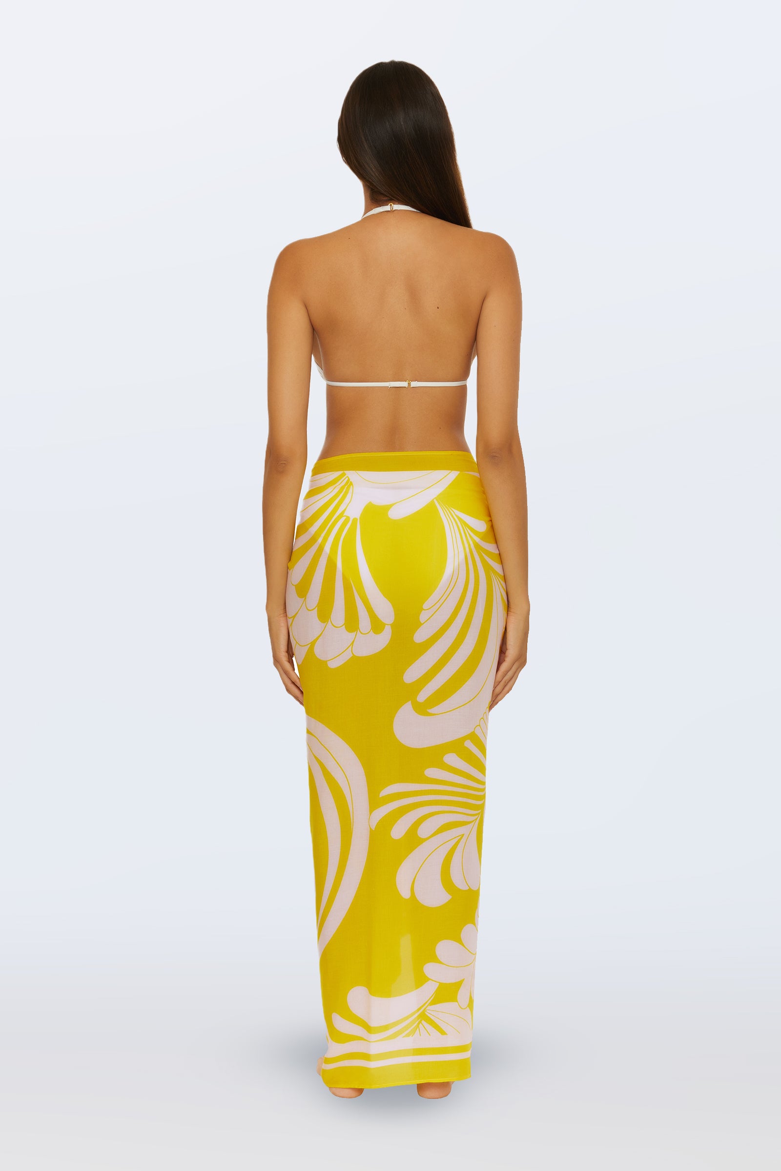 A woman faces away, wearing the Pareo Swim Cover-Up - Monaco Desert Sun—a long yellow skirt with bold white abstract patterns—paired with a white halter bikini top. Her straight brown hair is down against a plain white background.