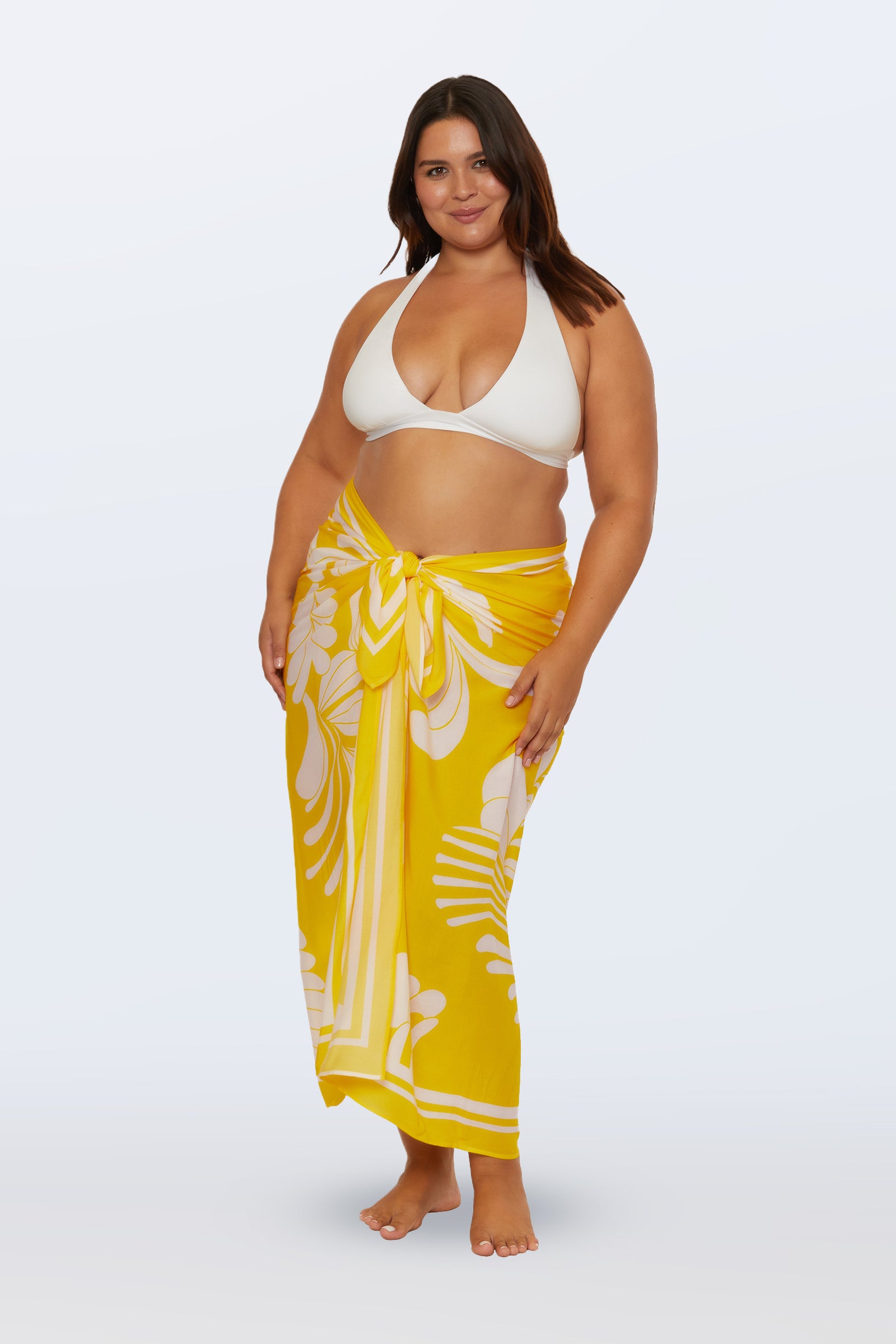 A woman wears the Monaco Desert Sun Pareo Swim Cover-Up, a yellow sarong with a white floral pattern, paired with a white bikini top. She stands barefoot against a plain light background, smiling with one hand on her hip.