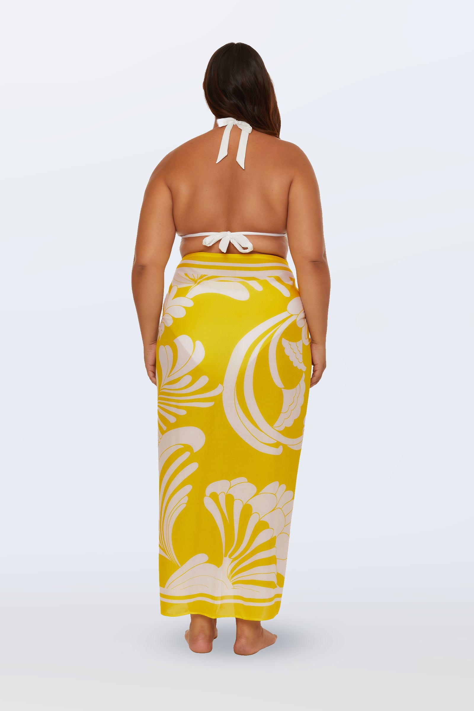 A woman stands barefoot with her back to the camera, modeling the Monaco Desert Sun Pareo Swim Cover-Up—a long yellow sarong with a bold white floral pattern—paired with a white halter bikini top against a plain, light background.