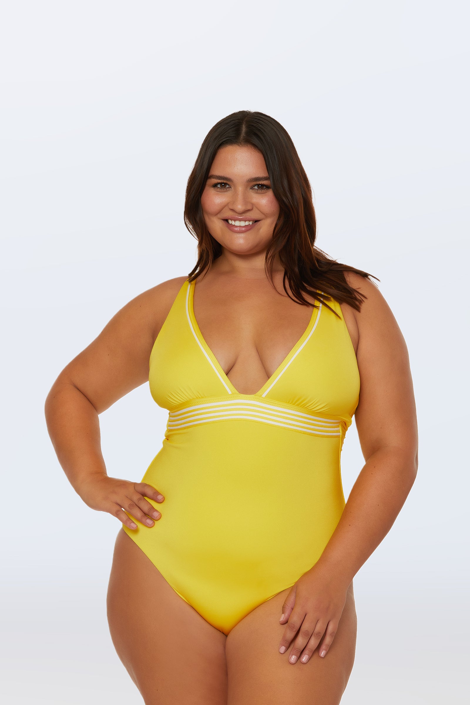 A woman with long dark hair smiles in the Monaco Pleated Plunge One-Piece, a bright yellow swimsuit featuring a plunging neckline and white striped details below the bust, standing against a simple light background.
