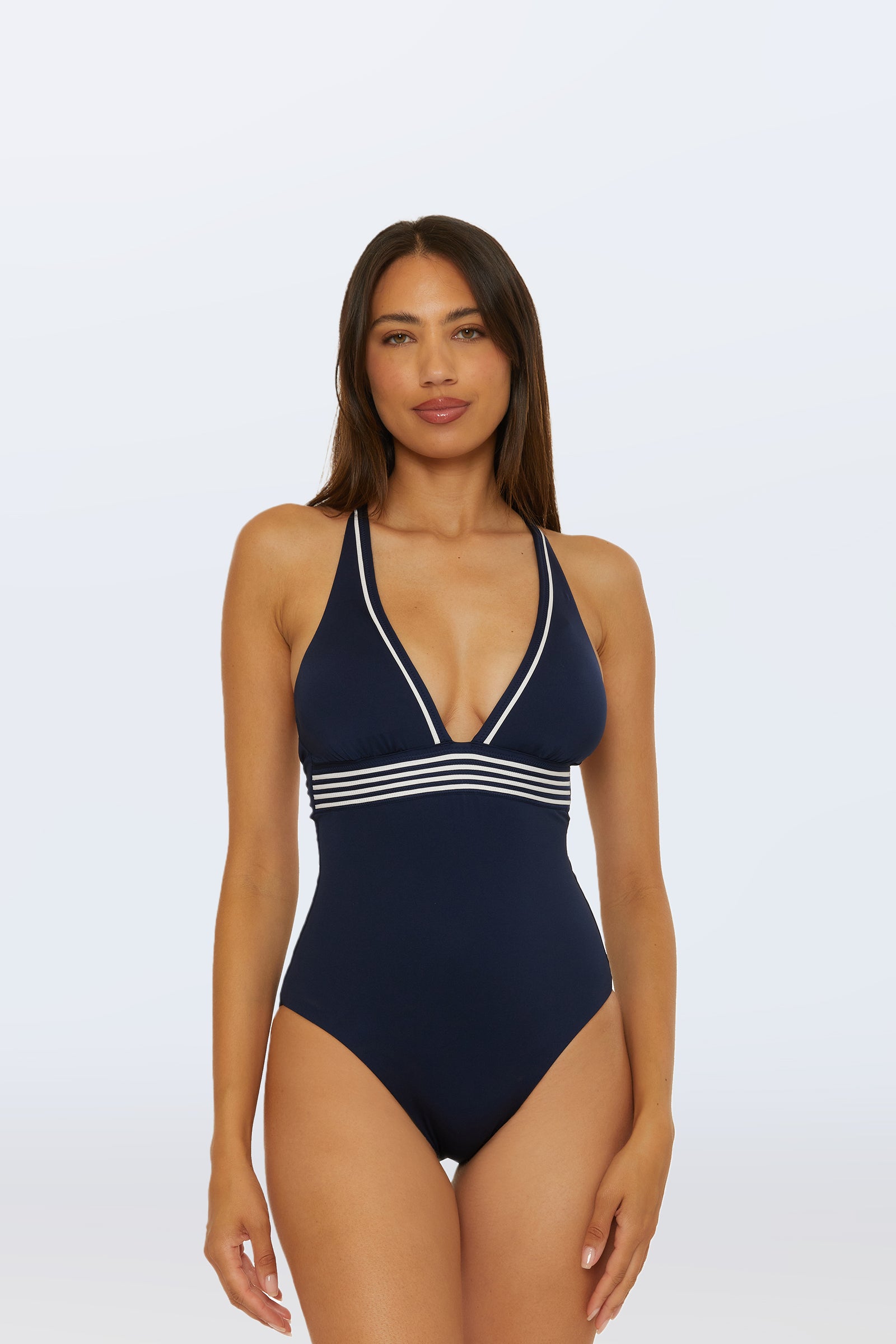 A woman models the Monaco Pleated Plunge One-Piece in Ink, featuring white trim and stripes under the bust, standing against a plain light background with a neutral expression.