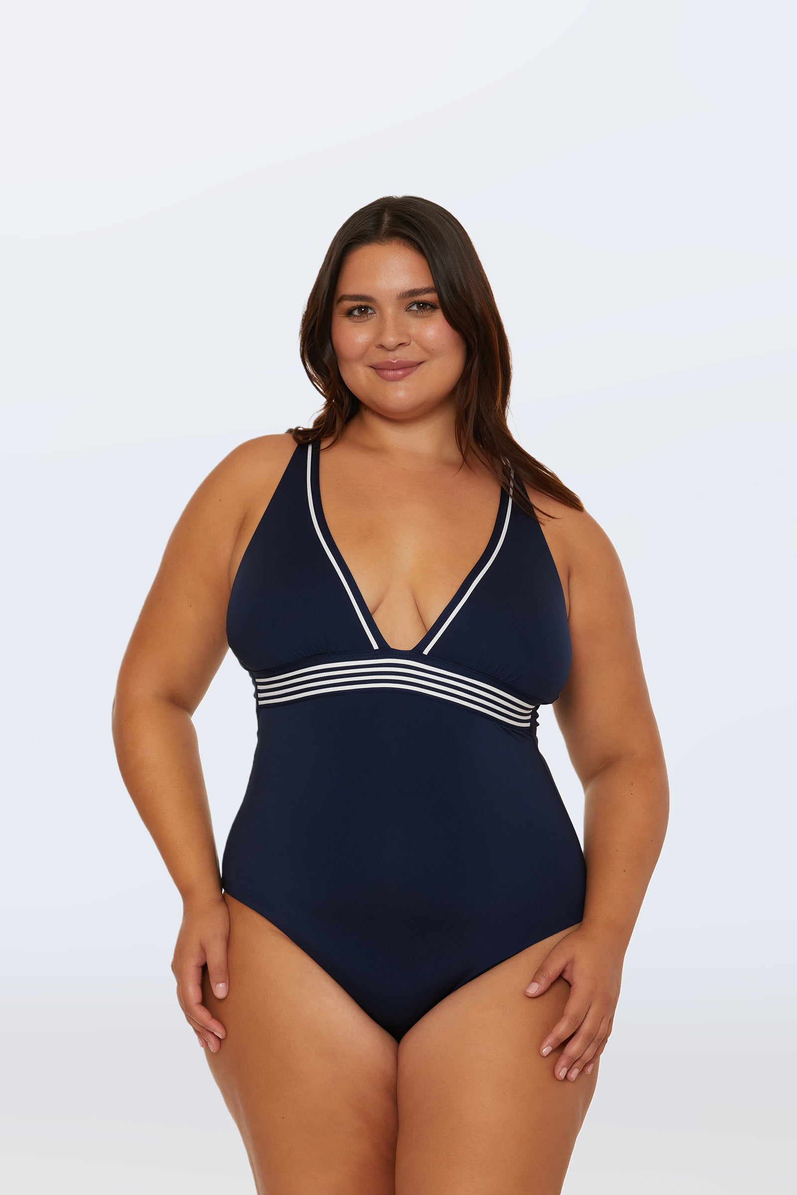 A woman with long brown hair smiles as she models the Monaco Pleated Plunge One-Piece, a navy swimsuit with a deep V-neck and white striped trim, standing against a plain light background.
