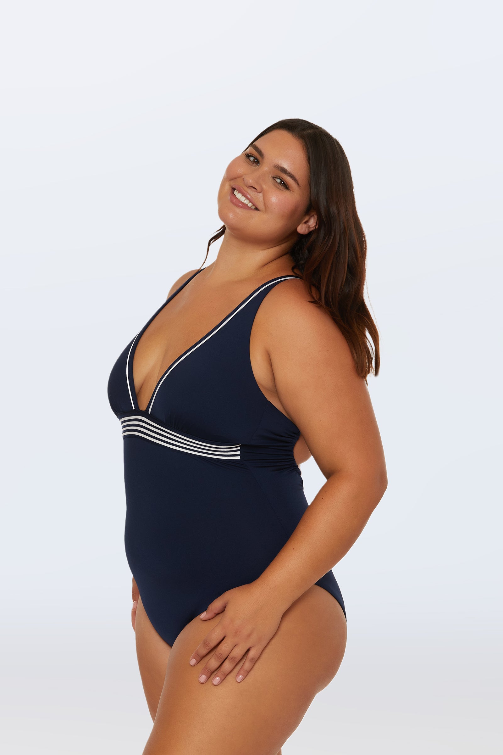 A woman with long brown hair smiles, modeling the Monaco Pleated Plunge One-Piece—a navy blue swimsuit with white trim and a deep V-neck—against a plain light background.