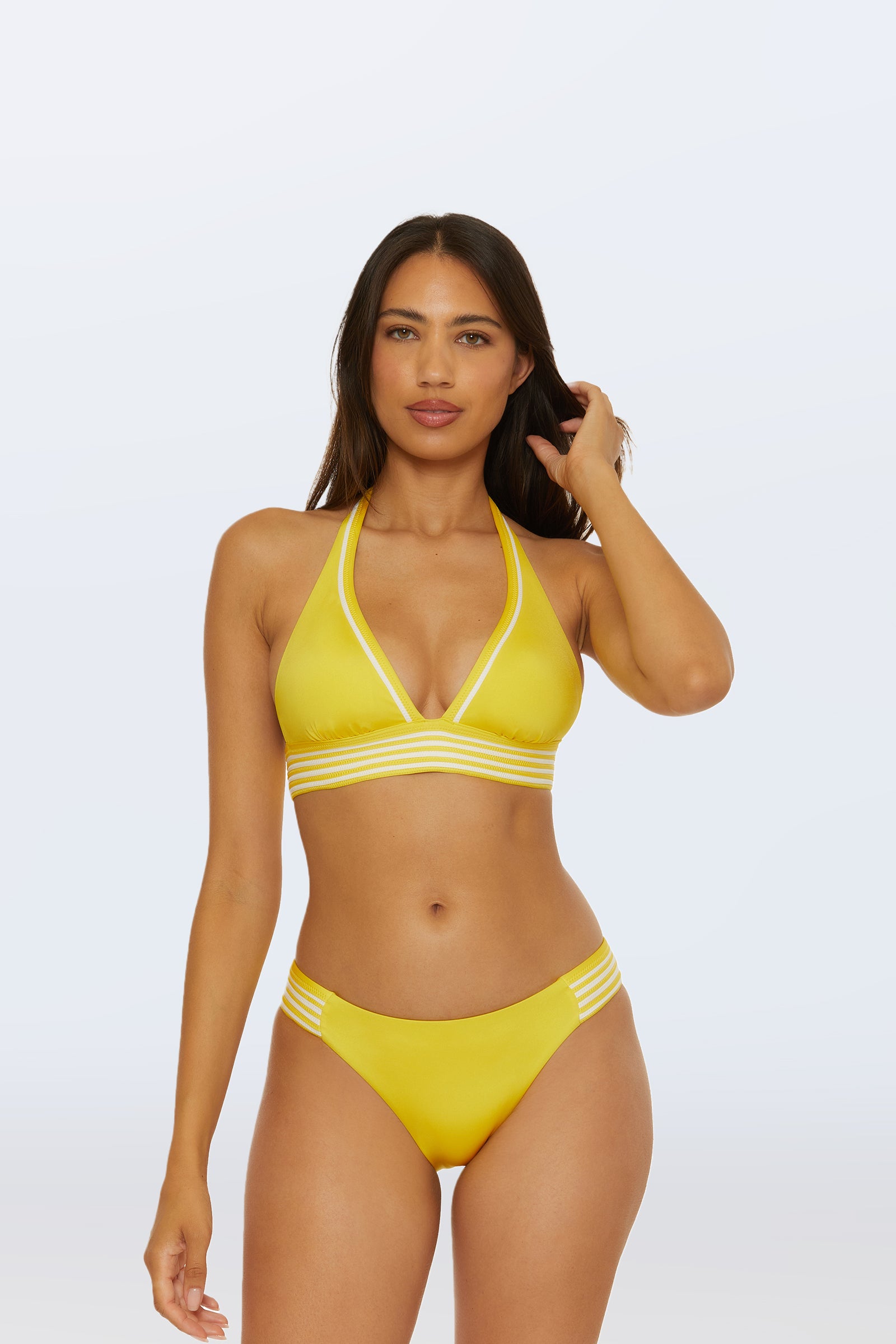 A woman models the Monaco Pleated Tab Hipster Bottom, a yellow bikini with white striped trim, standing against a plain light background and looking at the camera with one hand in her hair.
