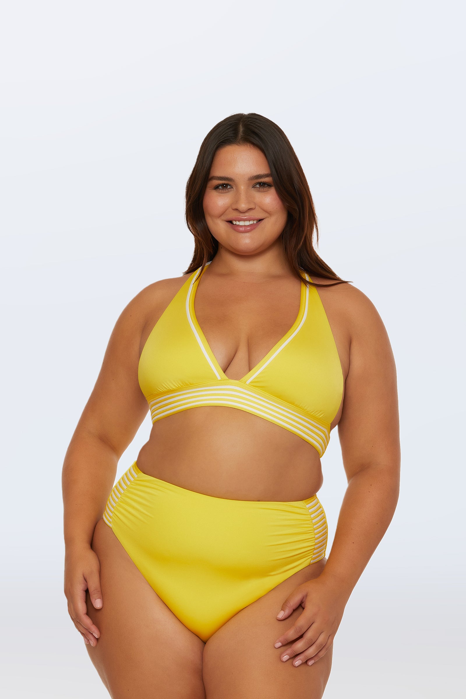 A woman with long brown hair smiles in the Monaco Pleated Halter Triangle Top, a bright yellow bikini with white and striped trim. The high-waisted bottoms have matching stripes. She stands against a plain light gray background.