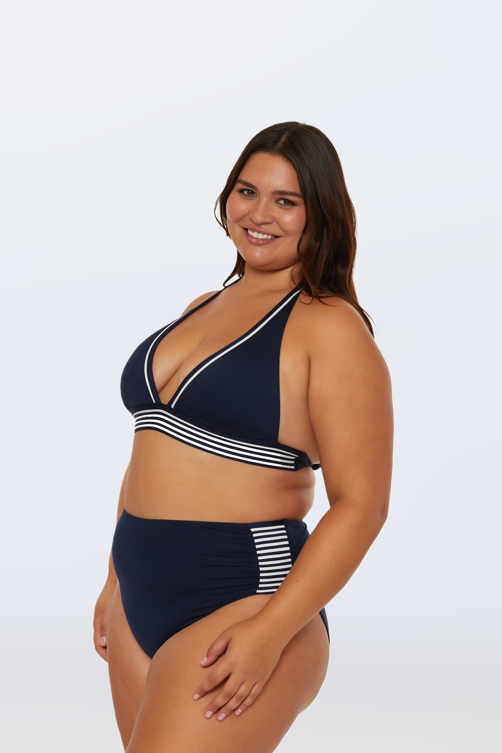 A woman with long brown hair smiles in a navy blue bikini featuring white striped trim and the Monaco Pleated High-Waist Bikini Bottom, standing against a light plain background.