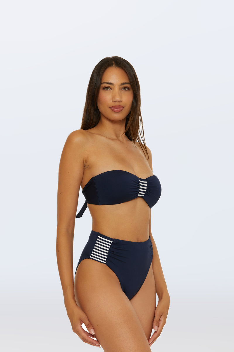 A woman with long brown hair models navy blue Monaco Pleated High-Waist Bikini Bottoms and a matching bandeau top with white stripes, standing against a light plain background.