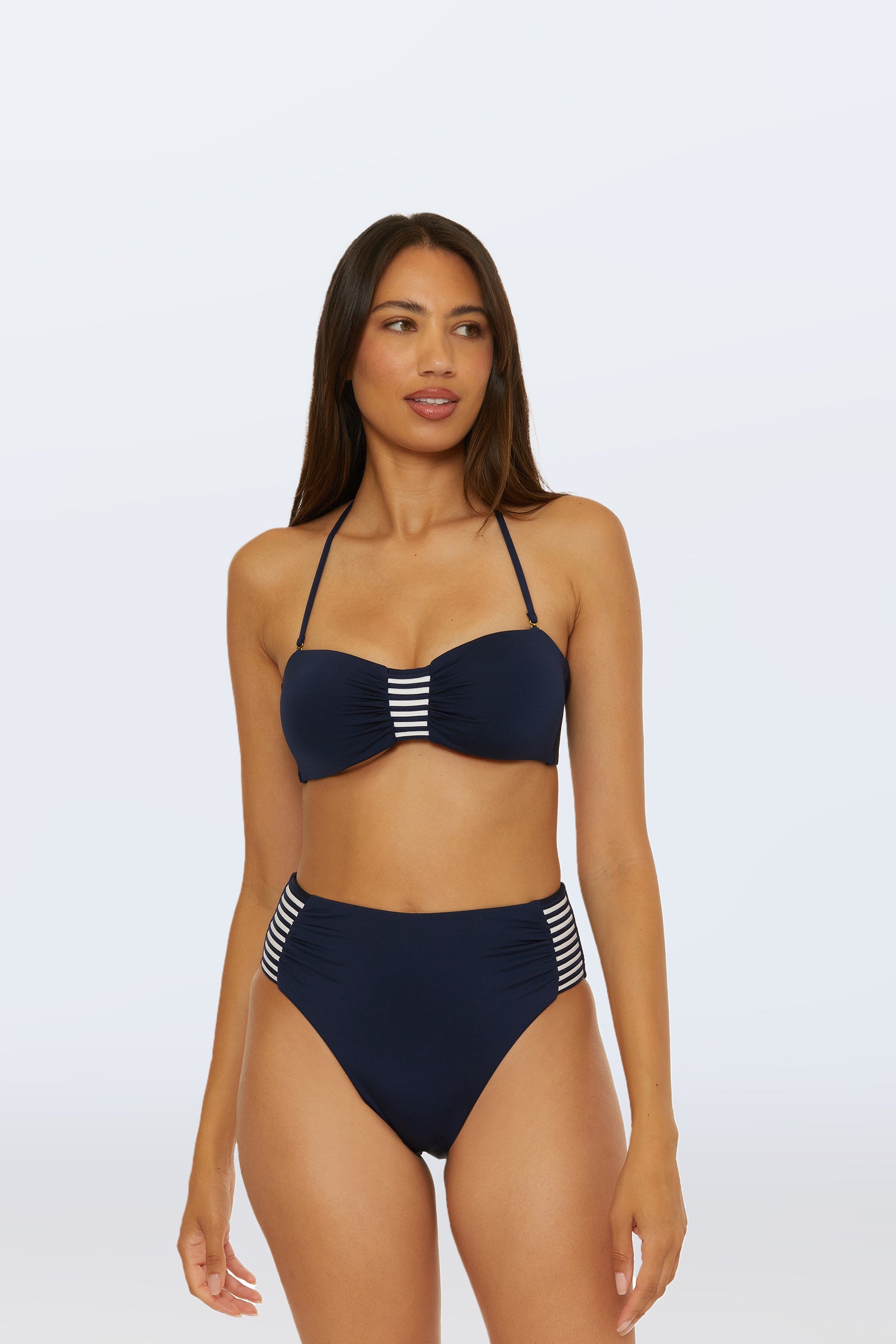 A woman models the Monaco Pleated Tab Bandeau Swim Top, a navy bikini featuring white stripes on the top center and sides of the high-waisted bottoms, against a plain light background.