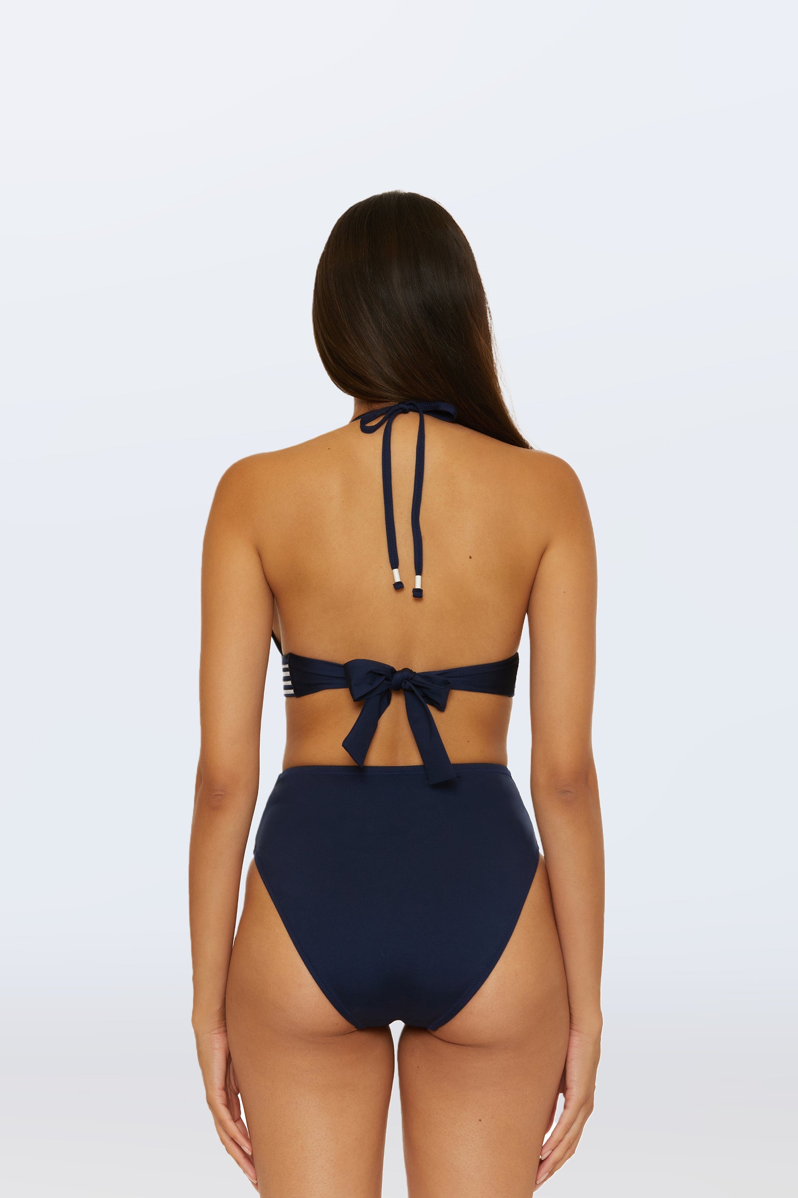 A woman with long brown hair is shown from the back wearing the Monaco Pleated Tab Bandeau Swim Top in navy blue, paired with matching high-waisted bikini bottoms, set against a plain light background.