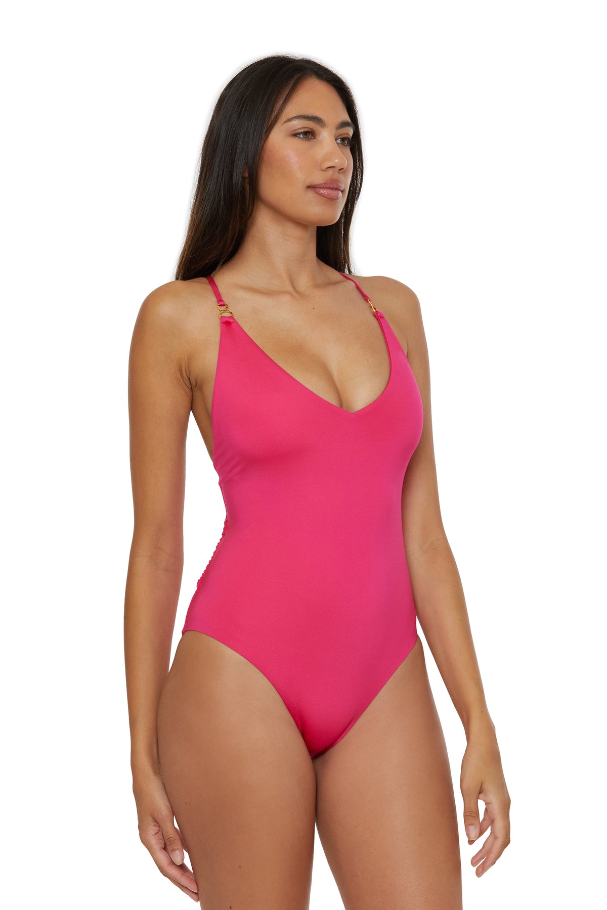 A woman with long dark hair wears the Monaco Twist Back One-Piece Swimsuit, a bright pink style with thin straps, standing against a plain white background and looking slightly to the side.