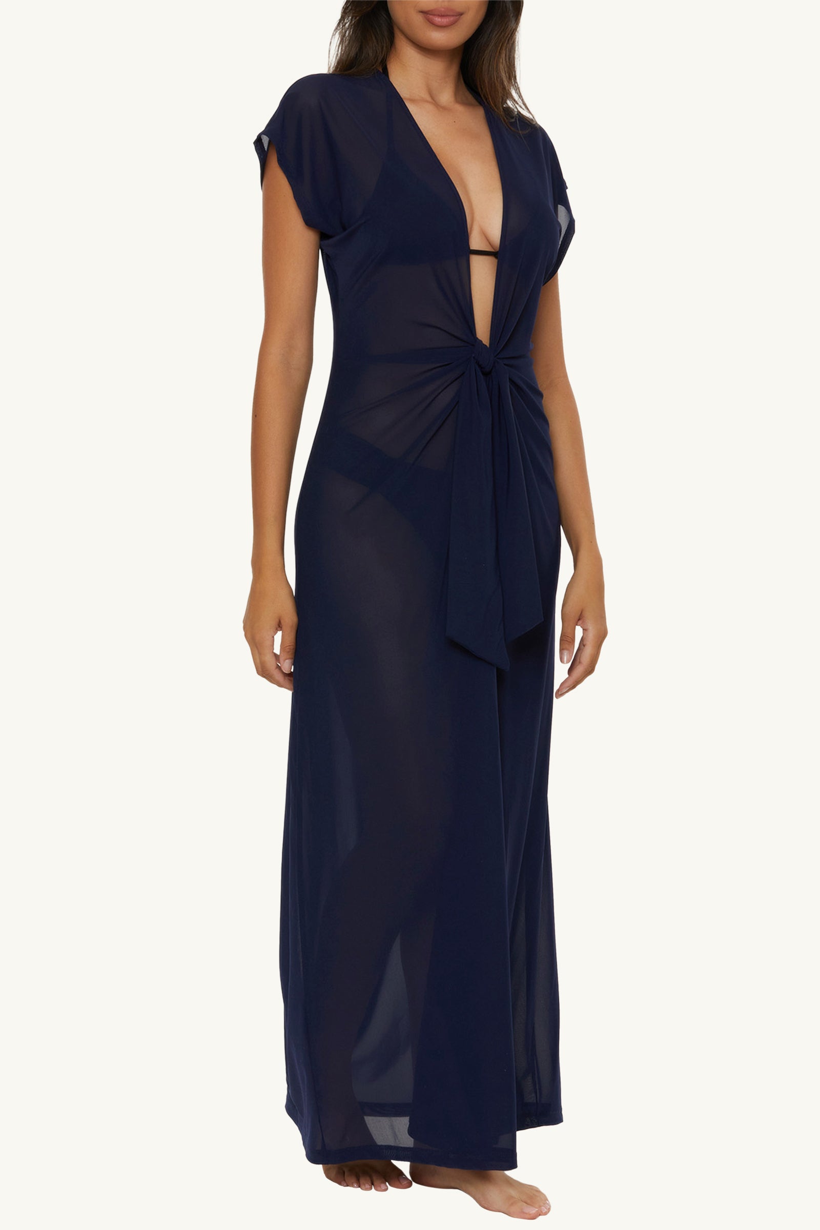 A woman wearing the Mesh Tie-Front Maxi Dress Cover-Up in Monaco, a sheer navy blue floor-length dress with short sleeves, deep V-neckline, and front knot detail, stands barefoot against a plain light background.