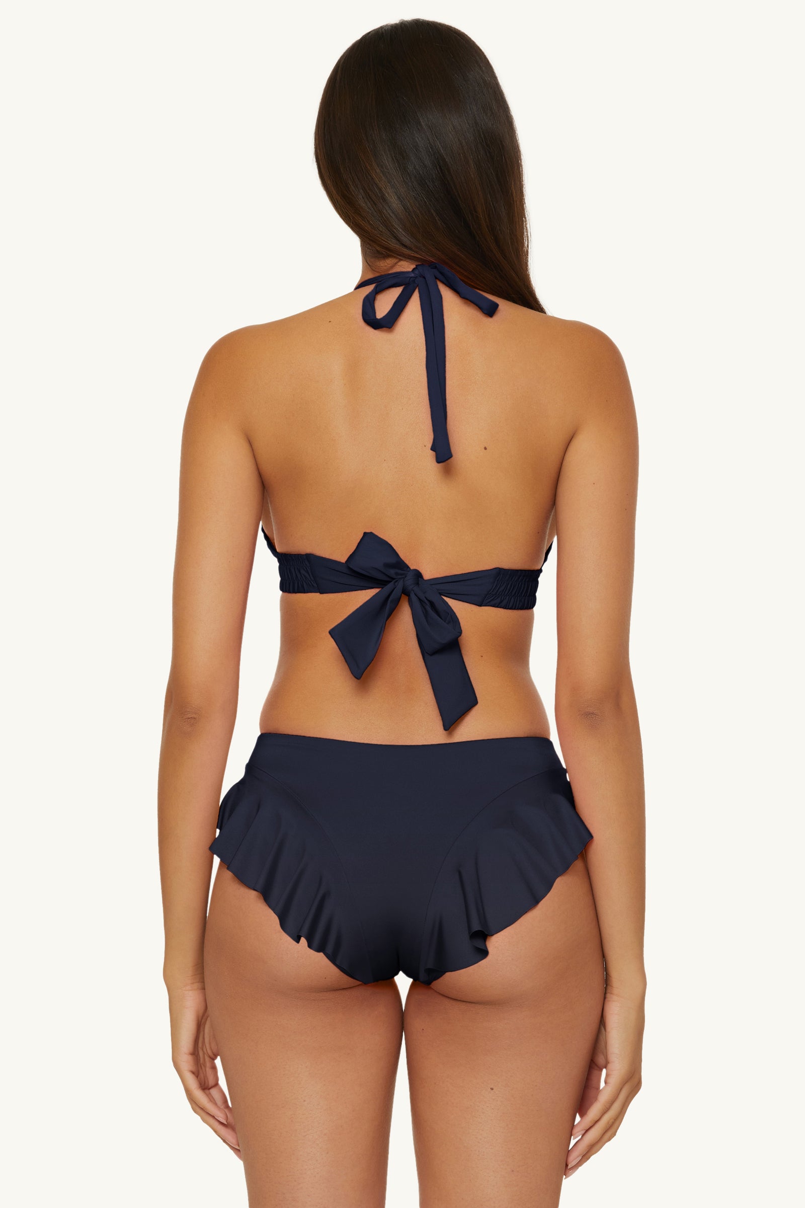 Woman with long brown hair is seen from behind, wearing an ink-colored halter bikini top and the Ruffle Hipster Swim Bottom - Monaco in Ink, featuring high-waisted ruffled details, against a plain background.